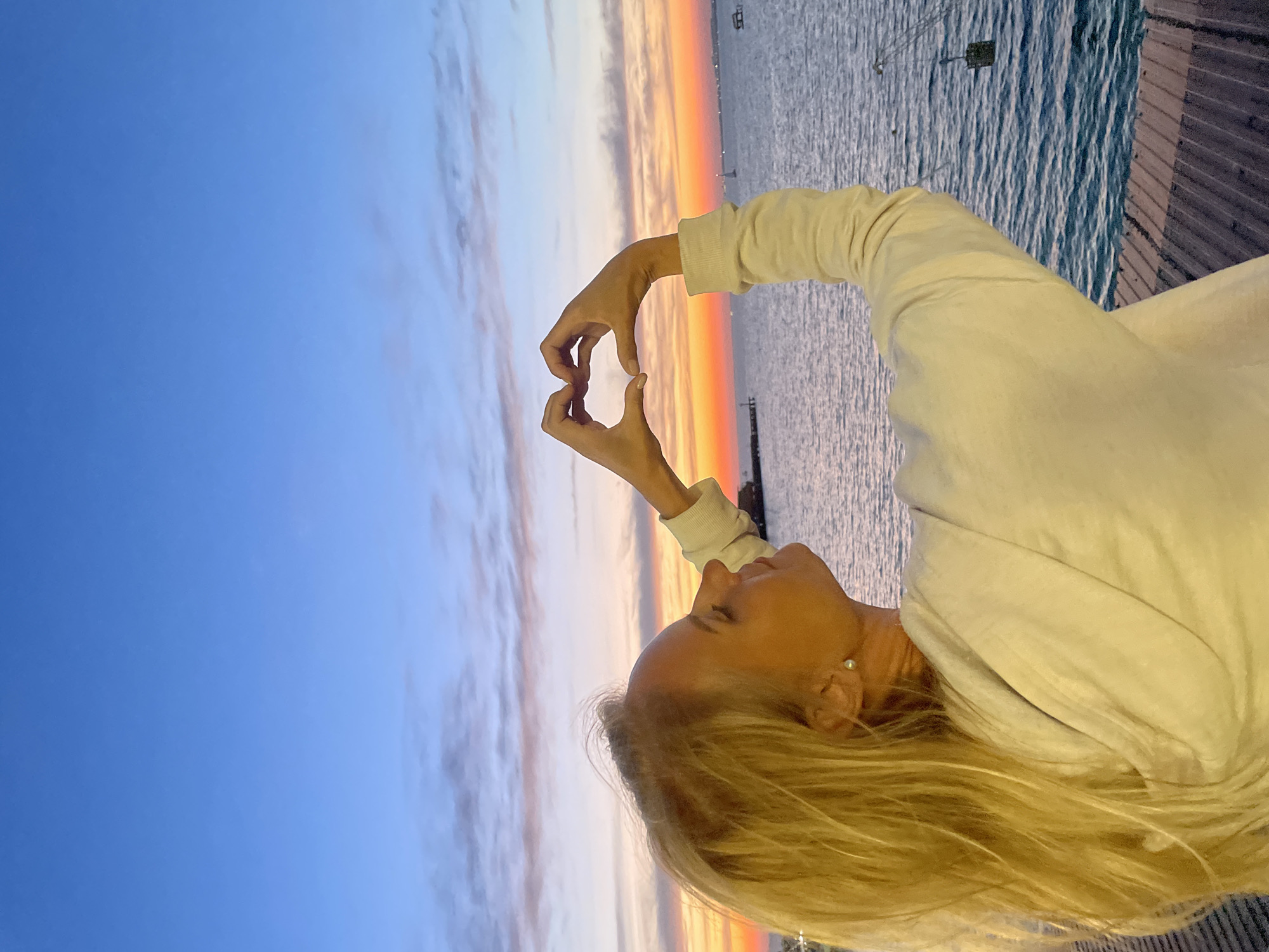A woman with blonde hair and a light-colored hoodie is standing on a wooden pier, forming a heart shape with her hands. She is looking up at a colorful sunset over the sea. The sky transitions from a deep blue at the top to shades of pink, orange, and yellow near the horizon. The sea is calm with some ripples. In the distance, there are faint structures, possibly piers or boats. The image appears to have been taken at dusk. The scene is located in Anapa, Russia.