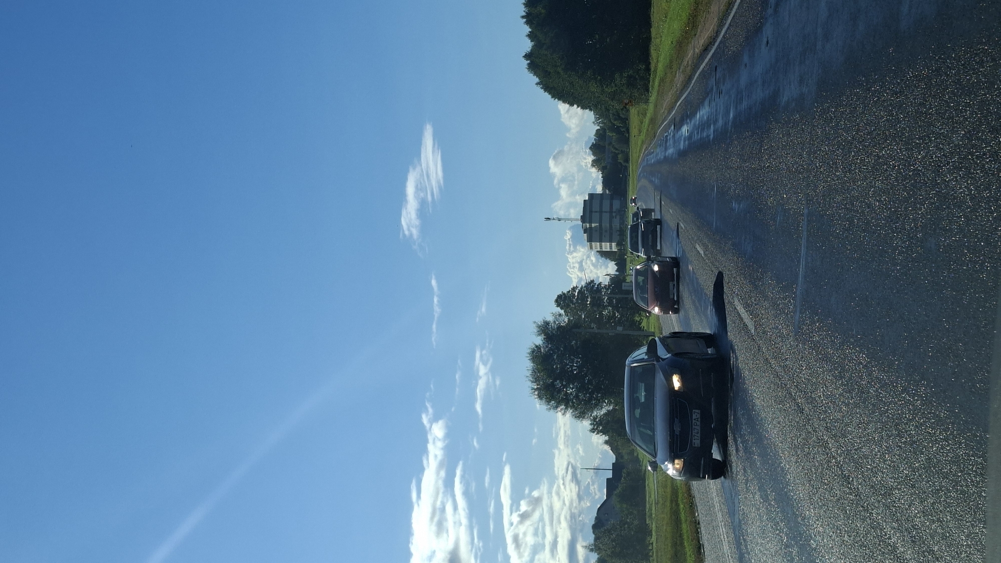 The image shows a view from a vehicle driving on a wet, asphalt road. The sky is clear blue with some scattered white clouds. Several cars are driving ahead on the road. In the background, there are trees on the sides of the road, and a multi-story building with a radio tower on top is visible in the distance. The license plate of the car in the foreground reads "1757 PA-7". The overall scene suggests a drive through a rural or suburban area on a sunny day.