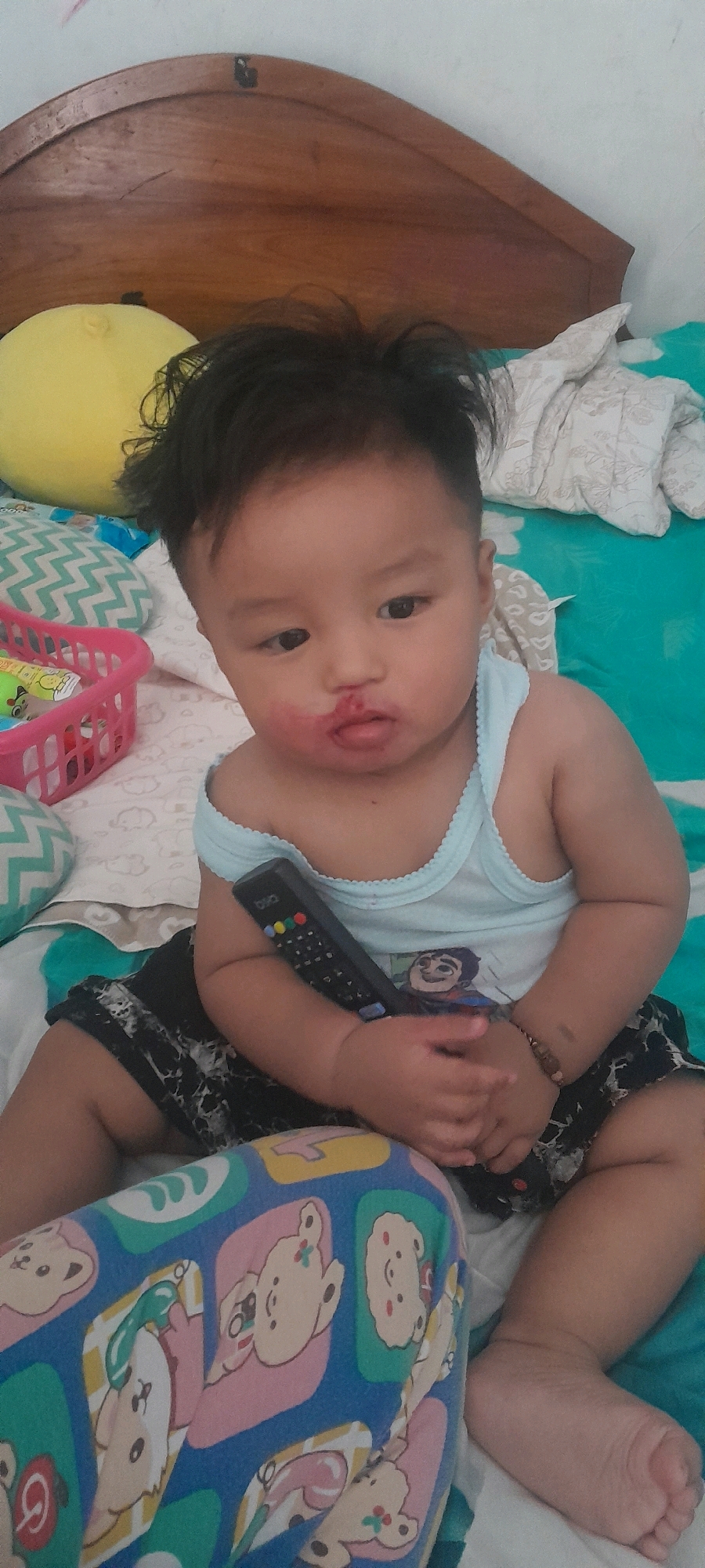 A toddler with dark hair and a smudge of red substance around their mouth sits on a bed. They are wearing a light blue tank top with a cartoon character on it and dark patterned shorts. The toddler holds a black remote control with colored buttons in their right hand. The background consists of a wooden headboard, pillows with geometric patterns, and a teal-colored sheet. A pink plastic basket is visible to the left of the toddler. The scene appears to be indoors, likely in a bedroom. There is no information available in the image to ascertain the specific location within the city of Sambong, Indonesia.