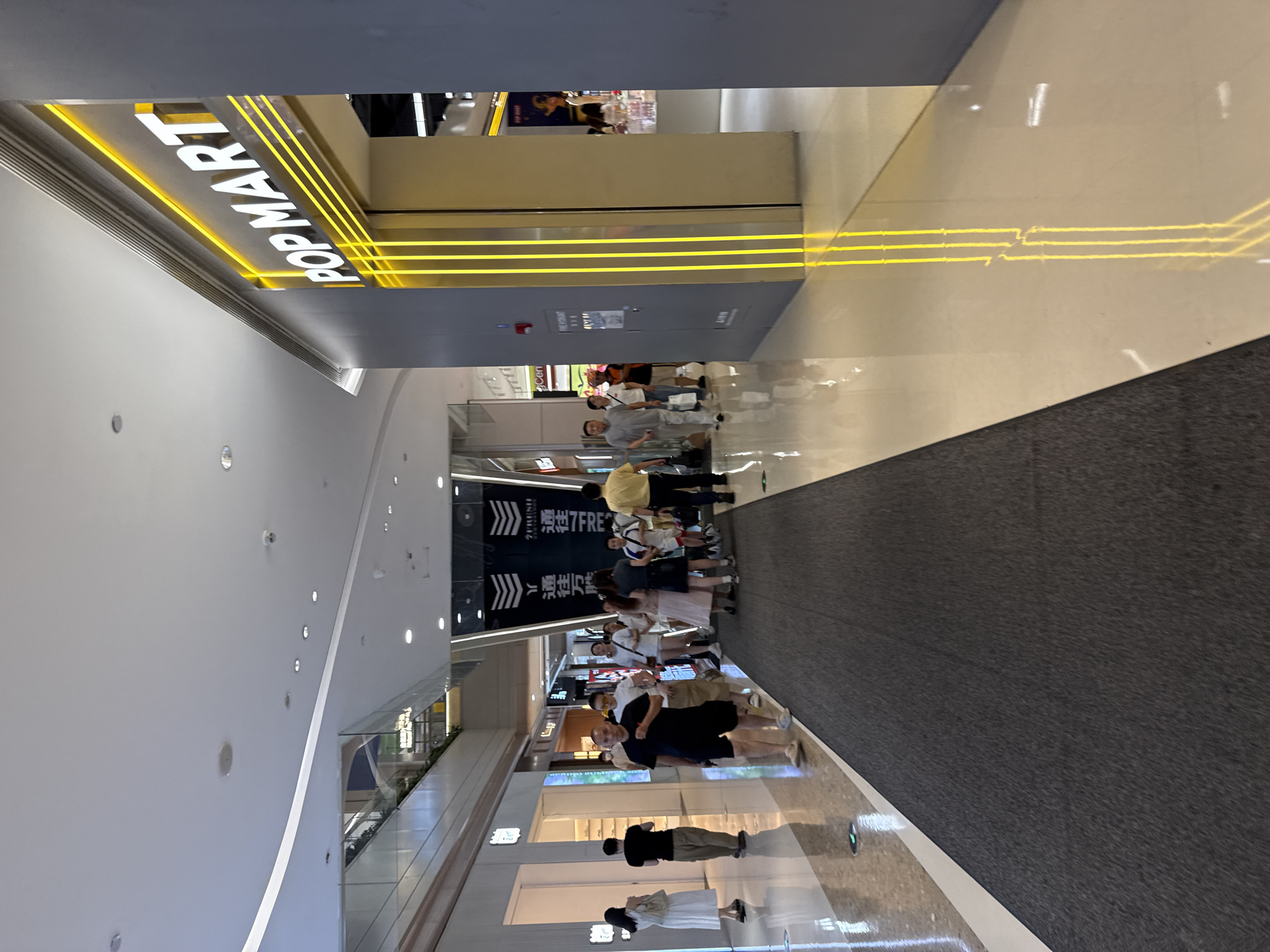 The image captures an interior view of a modern shopping mall, likely in Haizhu District, China. The foreground is dominated by a dark grey carpeted walkway that extends into the distance, bordered on the left by a polished light-colored floor reflecting overhead lights and store fronts. On the right, a prominent store called "POP MART" is visible, distinguished by its illuminated signage featuring white text on a dark background, framed by vertical yellow neon lights. Several people are seen walking along the carpeted pathway, with some exiting a store on the left and others moving towards the right. The architecture of the mall is characterized by clean lines, a high ceiling with recessed lighting, and large glass storefronts. The overall atmosphere suggests a busy retail environment.