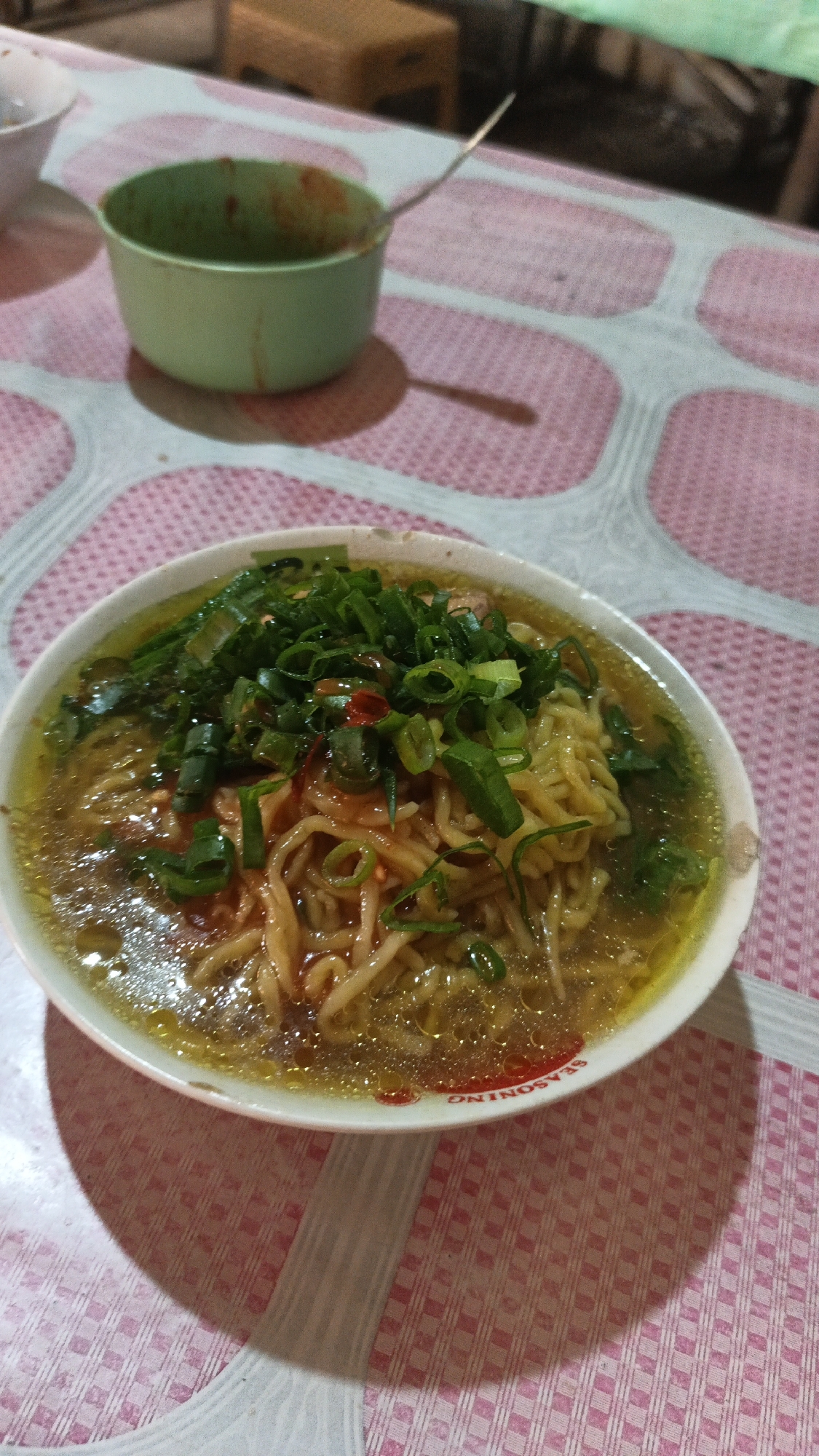 In Dawe, Indonesia, a close-up shot shows a bowl of noodle soup on a pink and white patterned tablecloth. The bowl contains ramen-style noodles in a clear broth, topped with chopped green onions and a few red chili flakes. To the left of the bowl, a small green dish with some red sauce and a spoon is visible. The background is out of focus, showing a hint of a wooden stool and what appears to be a table.