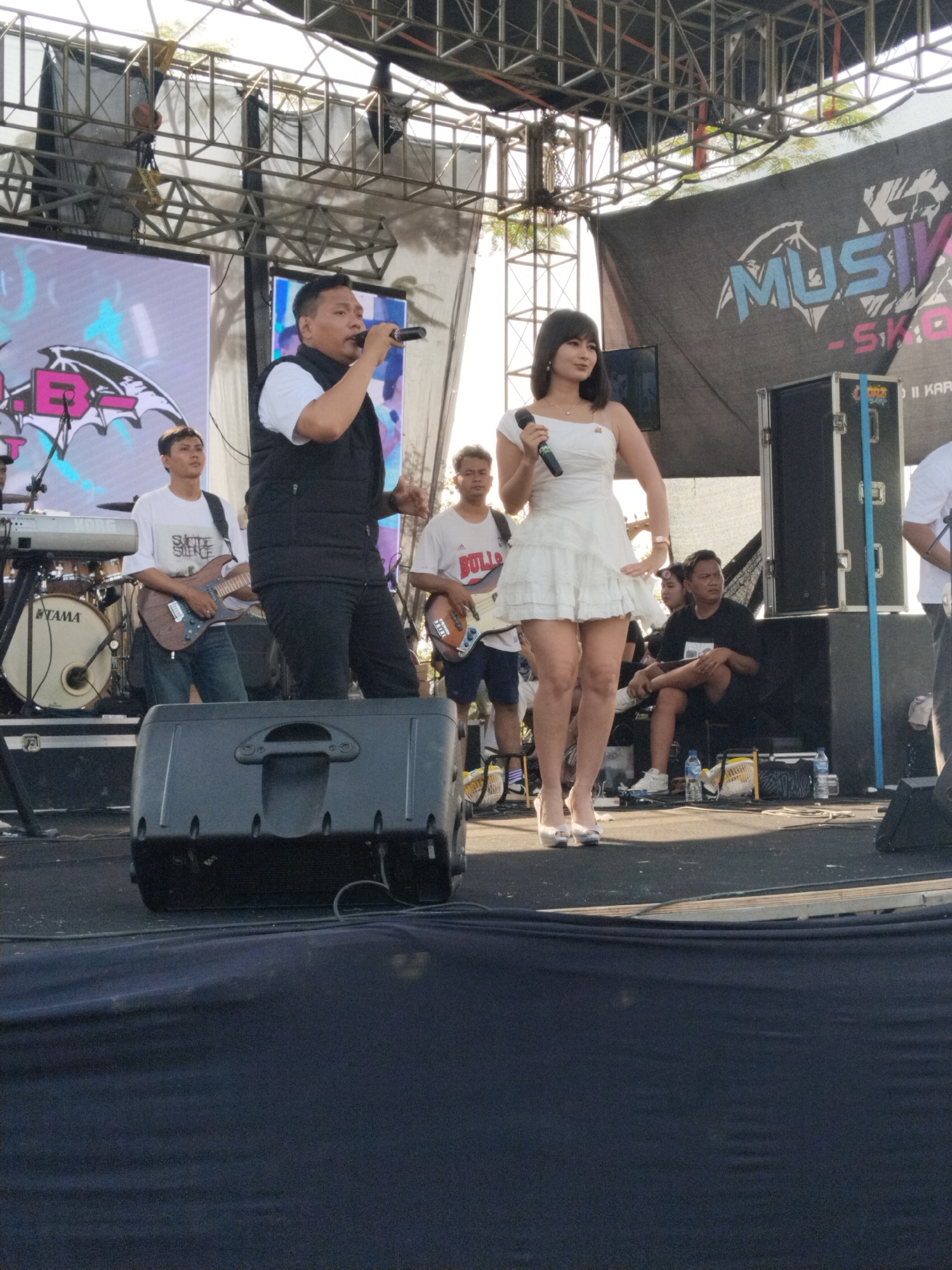 This image shows a stage with two singers performing. The male singer is wearing a white shirt, black vest, and black pants, holding a microphone and gesturing with his hand. The female singer is wearing a short white dress and silver high heels, also holding a microphone. Behind them are musicians playing instruments: a keyboard, a drum kit, and two guitars. A large screen is visible behind the stage, displaying graphics. The stage is set up outdoors, possibly for a music festival, in Karanganom, Indonesia. A banner with the word "MUSIKIVA" is also visible. A few people are seated or standing on the side of the stage. Sound equipment is located in front.