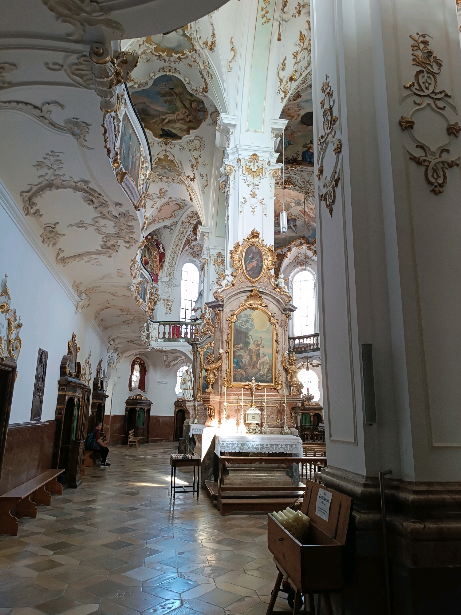 The image shows the interior of the Parish Church of St. Nicholas and St. Elizabeth in Andechs, Germany, a Baroque-style church known for its ornate decorations. The nave of the church is visible, with a high, elaborately decorated ceiling featuring frescoes and stucco work in white and gold. Sunlight streams through the windows, illuminating the tiled floor and casting shadows.
In the foreground, a wooden box filled with white candles sits on a stand. To the left, wooden pews line the nave, and a person is seated on one of them, facing towards the altar. The altar itself is a prominent feature, adorned with a large, ornate golden frame surrounding a painting. Behind the altar, a white tablecloth covers a surface, and a choir loft is visible above, with red railings. The walls are painted white and decorated with gold ornamentation. Confessionals with dark wood and green curtains are positioned along the left wall. The overall atmosphere is one of grandeur and spiritual serenity.