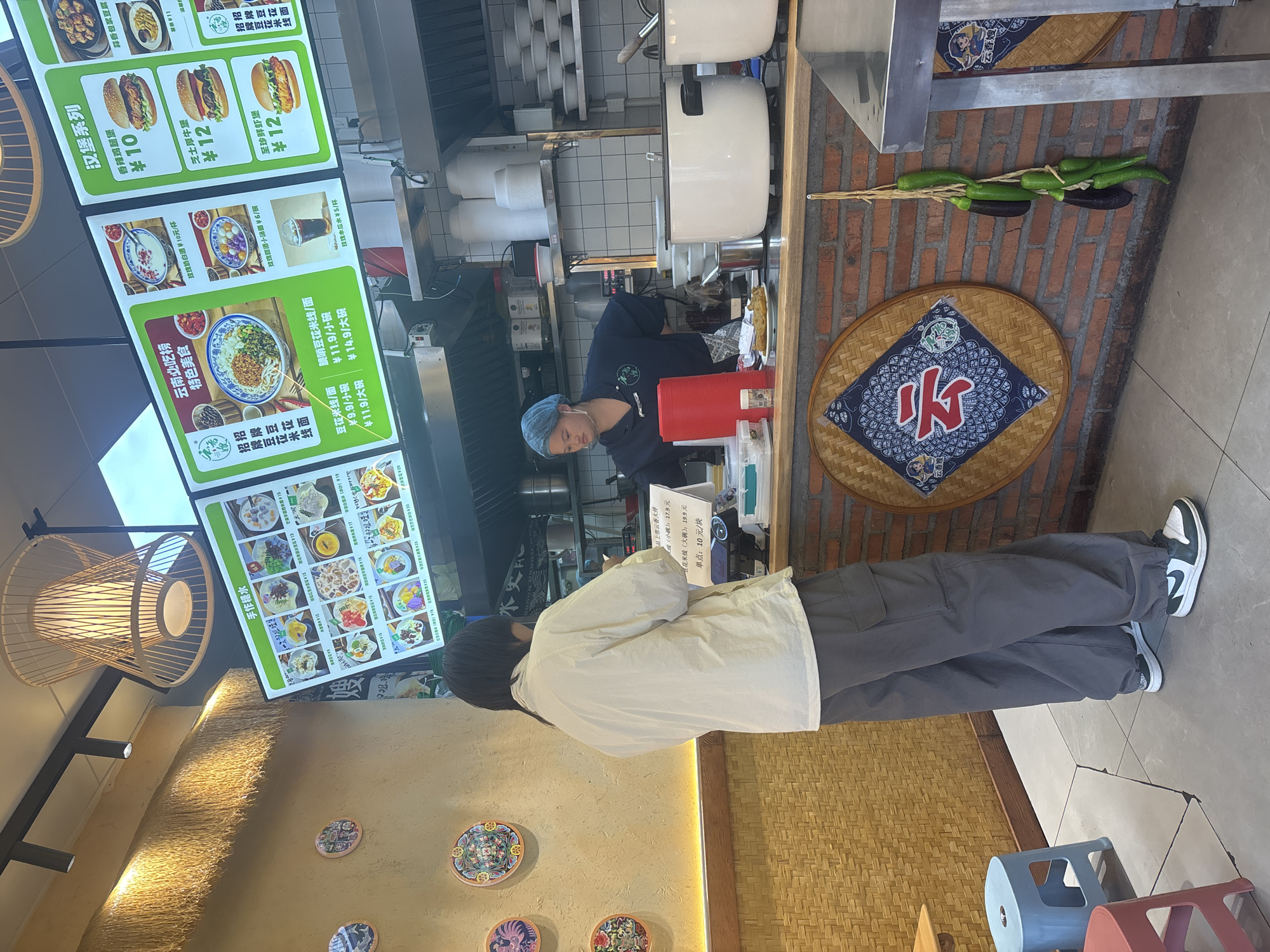 The image shows the interior of a restaurant in Suzhou Industrial Park, China. A person stands at the counter, looking at a menu displayed on a large digital screen. The menu features various food items, including noodles, burgers, and desserts, with prices listed in Chinese Yuan. Behind the counter, a person wearing a blue hairnet and apron works, with stacks of bowls and cooking equipment visible. Decorative items, such as woven placemats and hanging vegetables, adorn the walls. The restaurant has a casual, modern ambiance with elements of traditional Chinese decor.