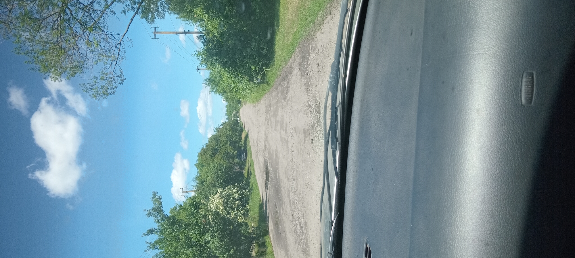The image, taken from inside a vehicle, shows a damaged asphalt road leading into the distance. The road is lined with dense green trees on both sides. Utility poles with overhead wires are visible along the right side of the road. The sky is blue with scattered white clouds. A portion of the car's dashboard, including the airbag cover, is visible in the foreground. The scene appears to be a rural or semi-rural area. The location is specified as Koviahy, Ukraine.