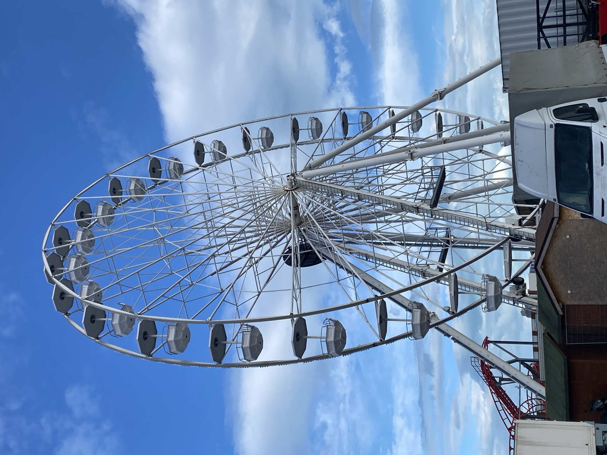 The image shows a Ferris wheel under a blue sky with scattered clouds. The wheel is primarily white, with evenly spaced gondolas or passenger cars around its perimeter. To the right of the wheel, there's a structure that includes a white van or truck and other metal elements, possibly part of the amusement park setup. The positioning of the items and vehicles near the wheel suggests the structures are part of the wheel’s supporting structure or related equipment for its operation. The image is probably taken during daytime. The location might be Varna, Bulgaria.