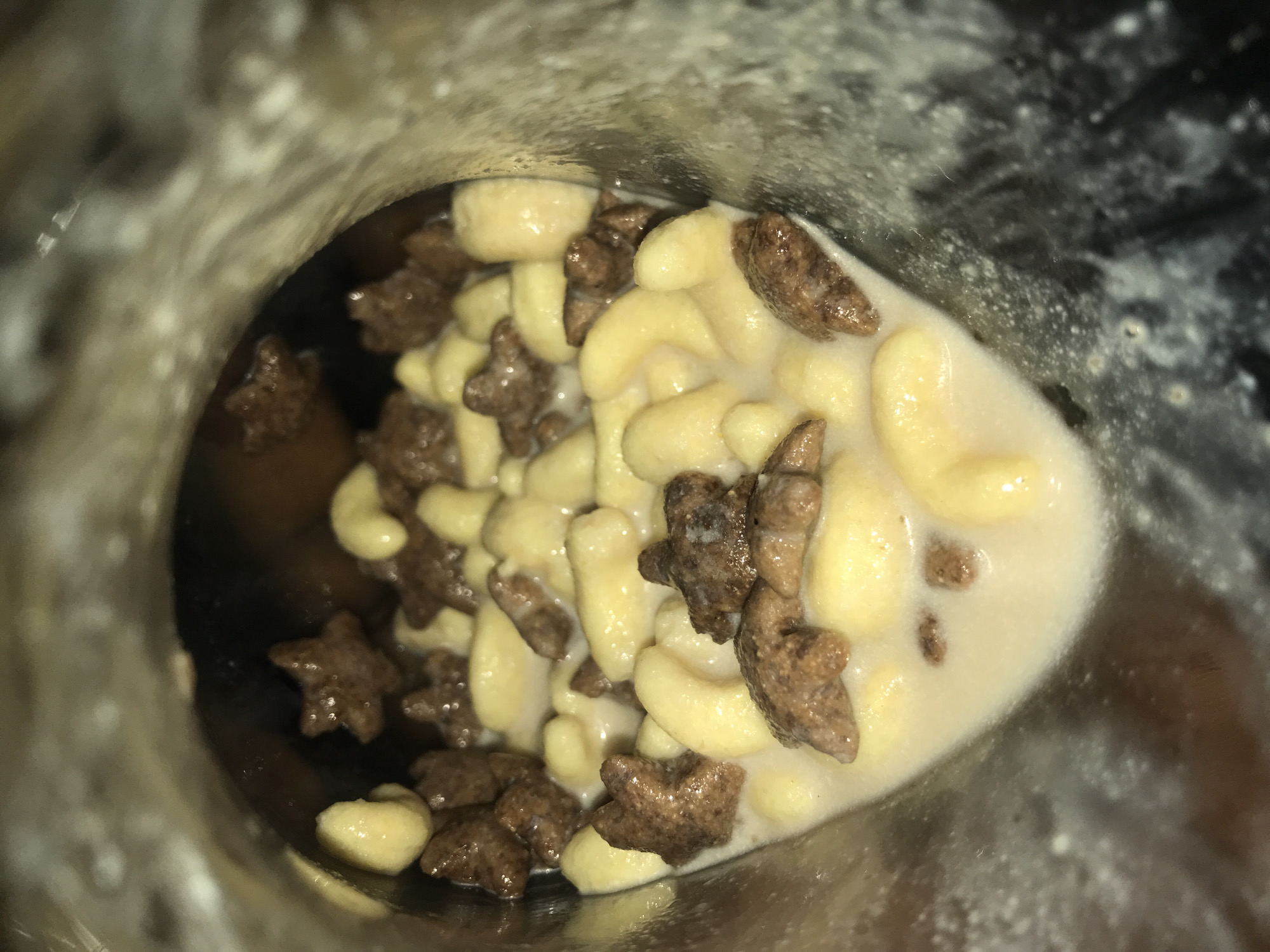 This image, captured in Rafin Par, Nigeria, depicts a close-up, downward view into the interior of a container, likely a bowl or mug. The container holds a quantity of milky white liquid partially submerging two distinct types of breakfast cereal. One cereal type is light yellow, presenting as elongated, curved, and tubular pieces. The second cereal type is dark brown, characterized by smaller, multi-pointed, star-like or irregular shapes. The cereal pieces are unevenly distributed within the liquid. The internal surface of the container appears light beige to brown, displaying some textural variations and scattered white residue. No individuals or dynamic actions are visible within the frame.