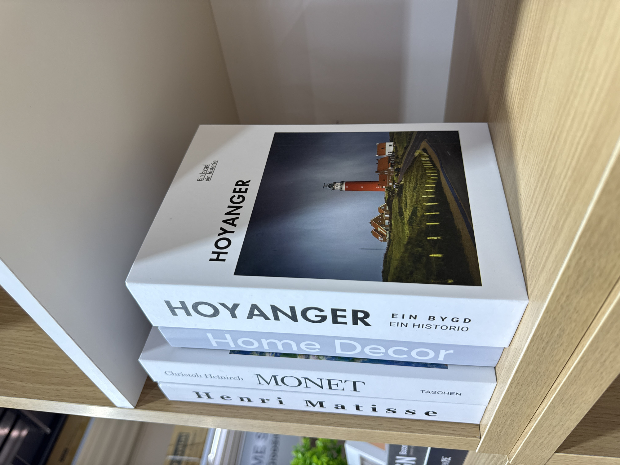 Four books are arranged on a light-colored wooden shelf. The books are white with text and images on their spines. From left to right, the visible text on the spines reads: "Henri Matisse", "MONET", "Home Decor", and "HOYANGER". The "HOYANGER" book has a large image of a lighthouse and houses on its cover, and the text "Ein bygd ein historio" is visible above the image. The background is a blurred interior space, possibly a store or home, with hints of other shelves and objects. There are no people or animals in the image. The scene is lit from the front, casting a slight shadow on the shelf. The overall impression is a clean and organized display of books. The location of Baan Chaweng Noi, Thailand is not discernible from the image.