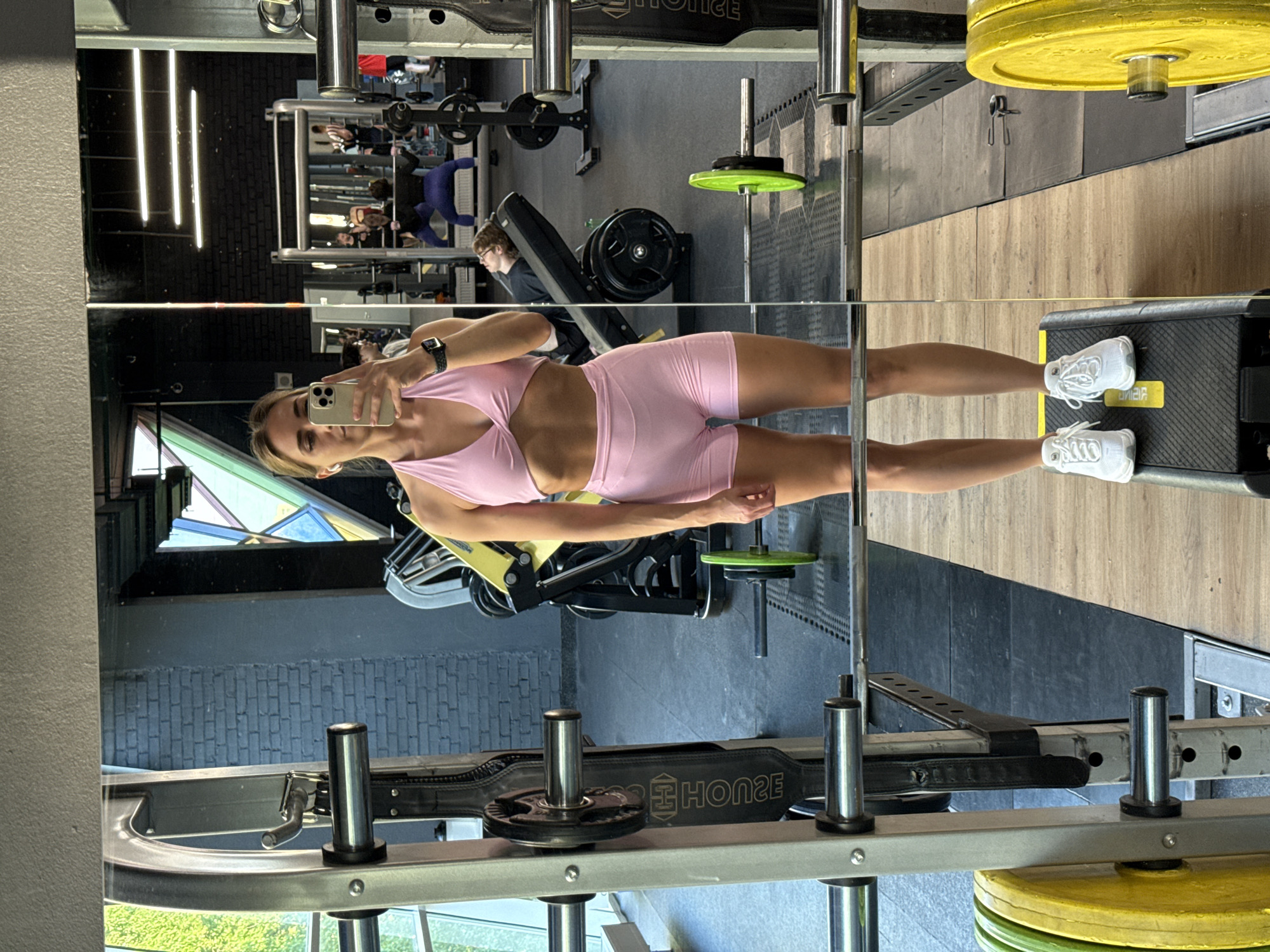 The image shows a woman in athletic wear taking a mirror selfie in a gym. She is standing on a black aerobic step with yellow accents, wearing a pink sports bra and pink shorts, and white athletic shoes. Her hair is blonde and pulled back. She is holding a smartphone in her right hand, angled to capture her reflection.
The gym environment is visible in the background through the large mirror. There are weightlifting machines, barbells with weight plates, and other gym equipment. In the reflection, other gym-goers can be seen. One person is on a weight bench, and at least two others are near weight racks. The walls appear to be a combination of dark brick and a textured gray material. Lighting fixtures are visible on the ceiling, and a large window with geometric framing is on the left side of the background, showing greenery outside.
The user has provided a location of Saint Petersburg, Russia, which is not visually verifiable from the image itself. The image is a full-body shot, focusing on the woman and her surroundings within the gym.