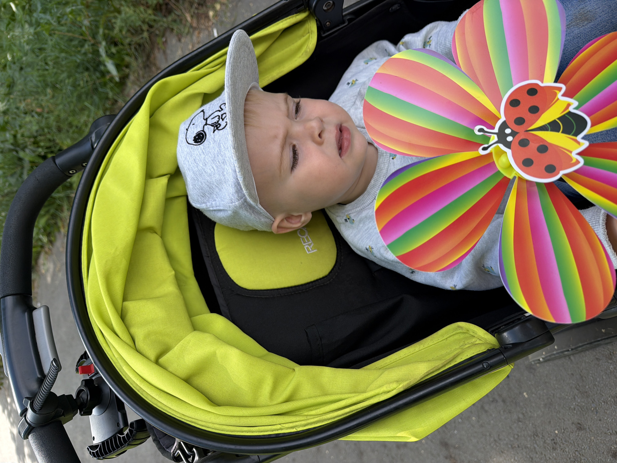 A young child with blond hair, wearing a grey baseball cap with a Snoopy graphic and a grey sweatshirt, sits in a lime green and black stroller. A colorful pinwheel toy with ladybug decoration is held in front of the child. The child's expression is one of slight displeasure. The stroller is positioned outdoors on a paved surface next to a grassy area with some foliage. The background suggests a park or residential street. There is no discernible connection to Votkinsk, Russia.