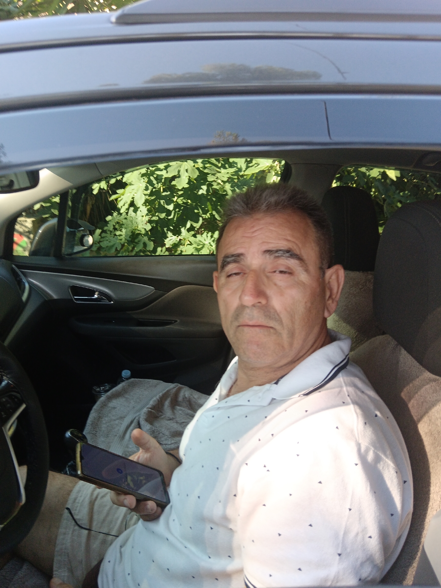 A man sits in the driver's seat of a car, looking towards the camera. He is wearing a white polo shirt with small dark triangular patterns and a blue and white striped collar. He is holding a smartphone in his right hand. The car interior is visible, including a portion of the steering wheel, the dashboard, and the passenger seat. Green foliage is visible through the car window, suggesting an outdoor setting. The lighting indicates it is daytime. No specific details about the "Municipal Unit of Vocha, Greece" are discernible from the image itself.