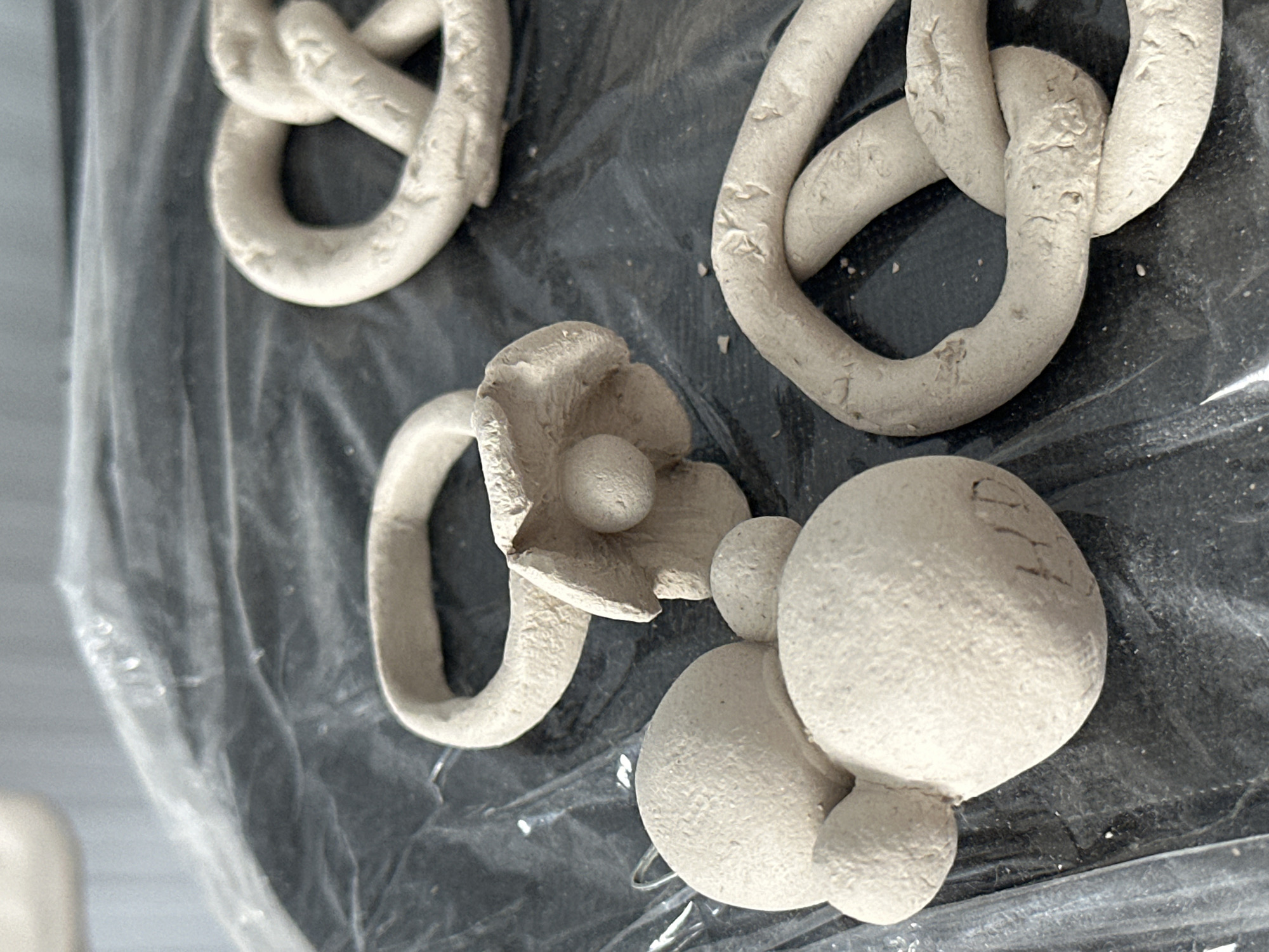 The image displays several unfinished clay sculptures on a black plastic surface. In the foreground to the left, there is a sculpture resembling Mickey Mouse, with a large circular head and two smaller circular ears. Engraved on the side of the larger circle are the letters "HD". To the right of the Mickey Mouse sculpture is a ring with a flower-like embellishment. In the background, on the right side of the frame, are two sculptures of pretzels. The overall scene is indoors, with a soft, diffused light source. There are no people or any signs of activity or interaction. The environment appears to be a crafting or art space. The specific location in the city of Nevinnomyssk, Russia, cannot be determined from the visual information provided.