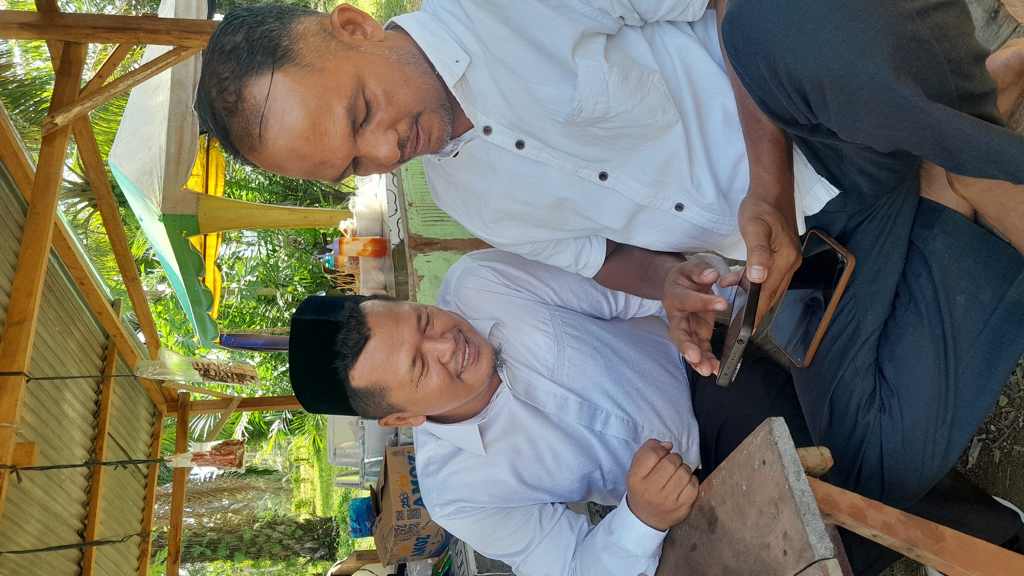 The image depicts two men in Blangriek, Indonesia, sitting outdoors under a wooden structure with a corrugated metal roof. The men are both wearing white shirts; one also wears a black peci (cap). They are looking at a cell phone. One man is holding a second cell phone. The environment is lush with vegetation. A box of AMOZ brand items is visible, partially obscured.