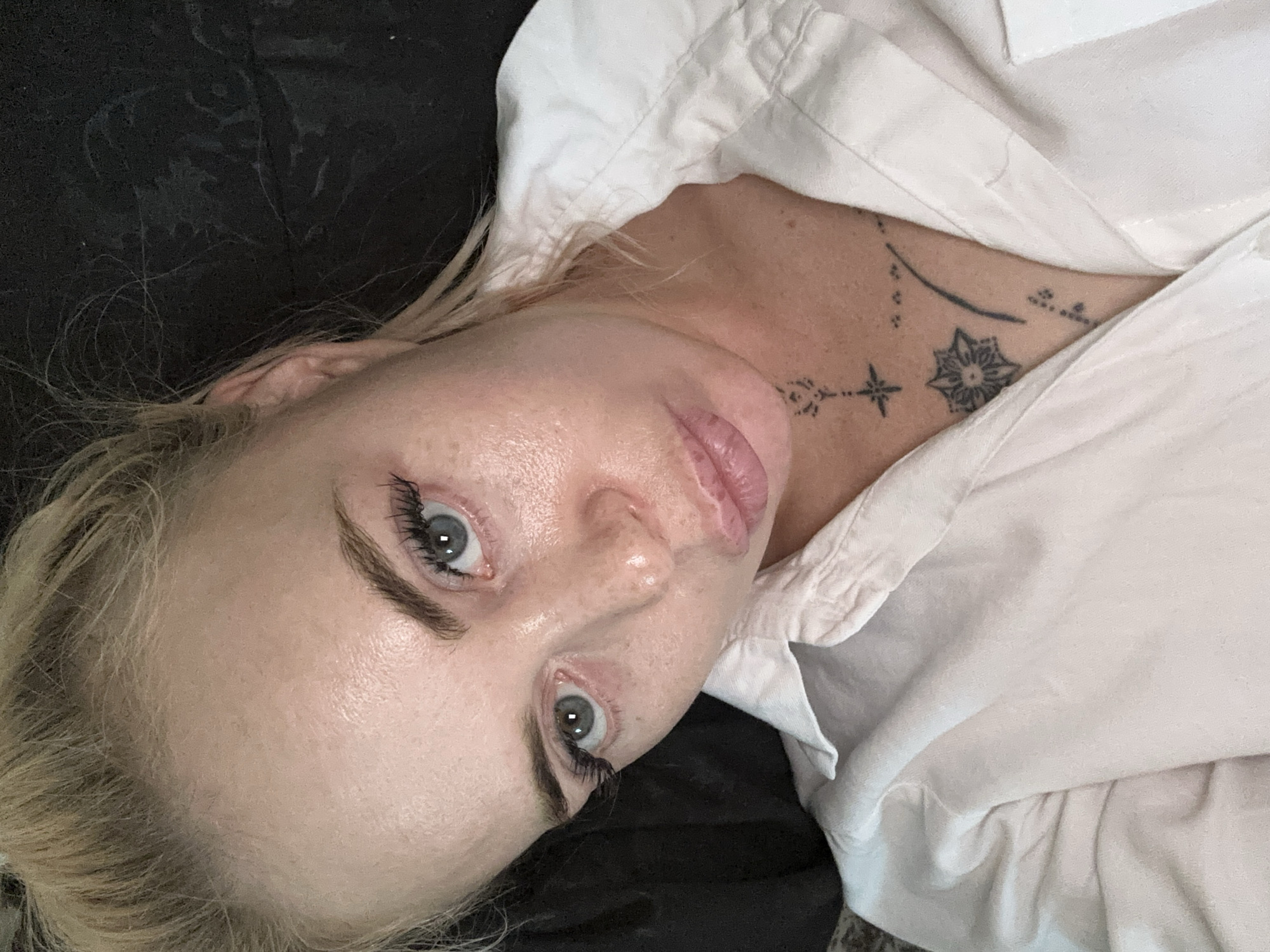 A close-up selfie of a blonde woman with blue eyes and full lips, wearing a white button-down shirt, is angled toward the viewer. Her hair is pulled back, revealing light-colored skin with some texture and faint freckles. She is wearing eye makeup, including mascara and false eyelashes. On her chest, several small black tattoos are visible, including a star and a floral design. The background is dark and out of focus, suggesting an indoor setting. The lighting appears to be from above and slightly to the side.