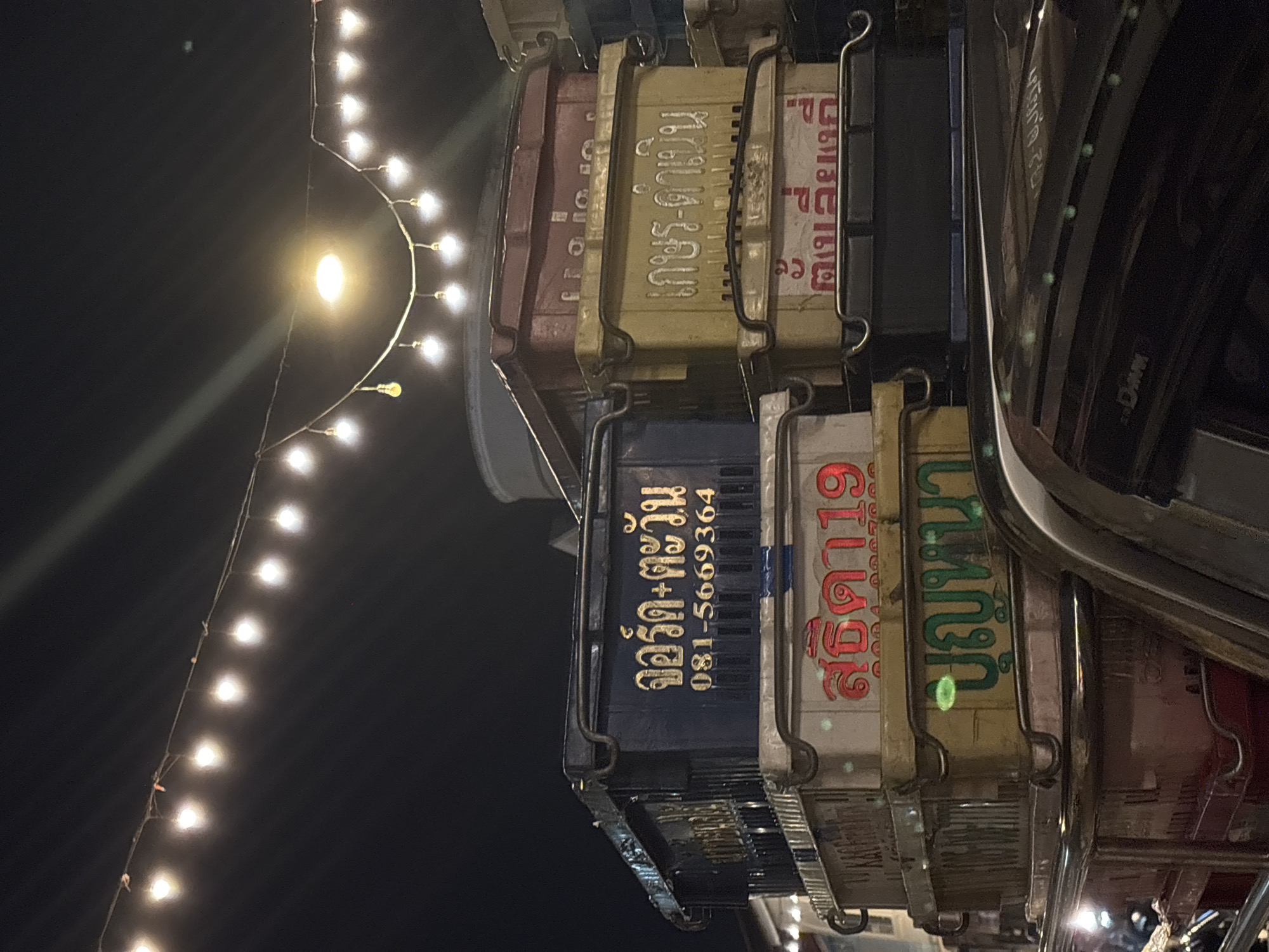 The image shows a close-up, low-angle shot of a vehicle carrying several stacked crates with Thai writing on them, with a string of lights visible in the background, likely taken at night in the Phuket City Municipality, Thailand. The crates are various colors including brown, yellow, blue, and white with lettering in Thai. The string of lights is curved above the crates, providing illumination. The vehicle's chrome frame is visible on the right. The Thai writing on the crates include, "จอร์ก-ตะวัน" (Jorg-ตะวัน) along with a phone number "081-5669364" and "เกษร-ดาเป็น" (Ketsorn-Da Pen).