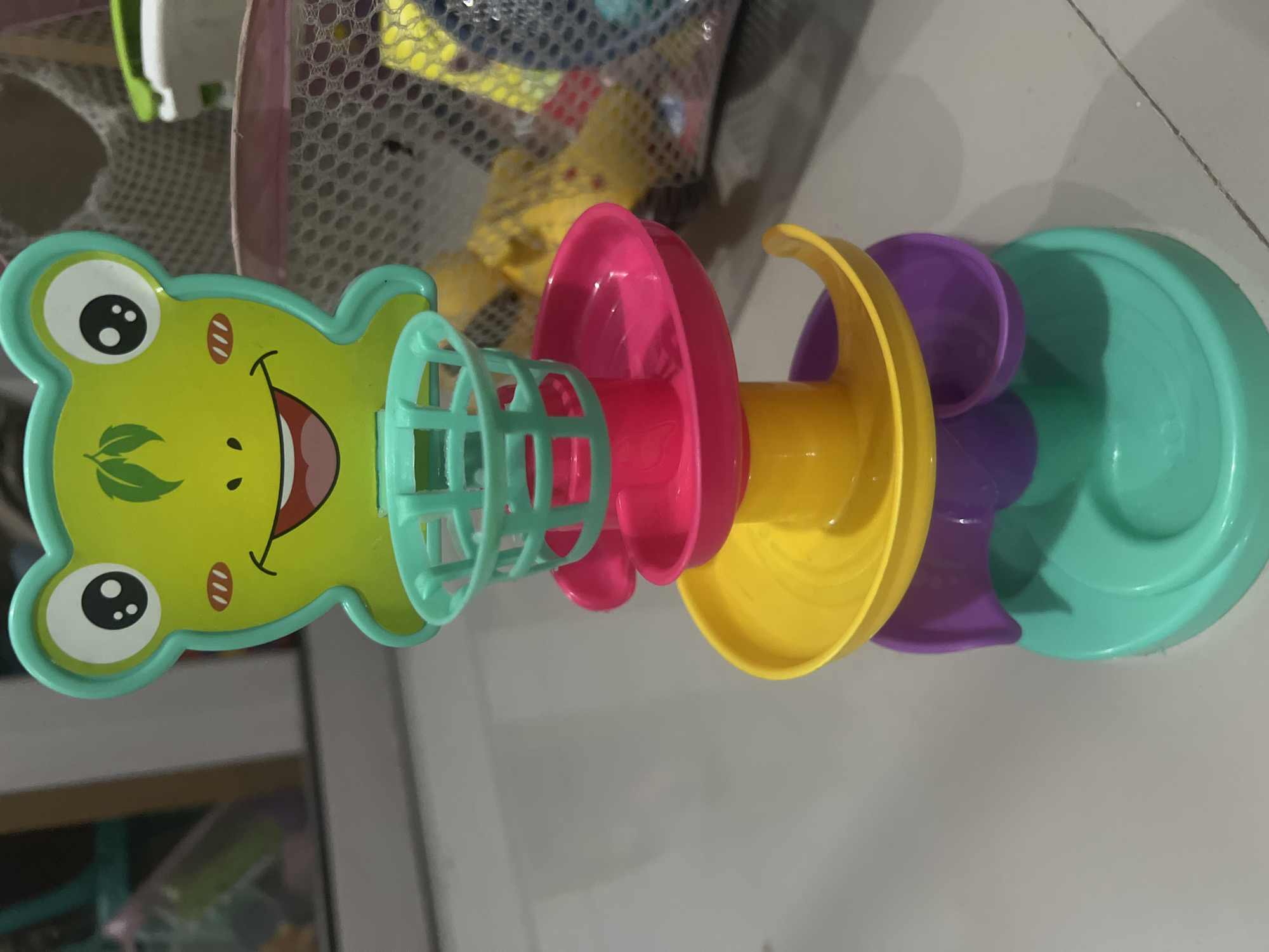 A frog-shaped toy with a basketball hoop sits on a tiled floor. The toy is green with a smiling face, big eyes, and rosy cheeks. Below the frog, the toy has a tower of colorful tracks and cups, including pink, yellow, purple, and teal. The toy is positioned in what appears to be a domestic indoor setting, possibly a child's playroom or living area, with blurred background elements suggesting furniture and possibly other toys in a net bag. The lighting is diffused, casting soft shadows. There is no indication of the city of Klapanunggal, Indonesia, within the visual information provided.