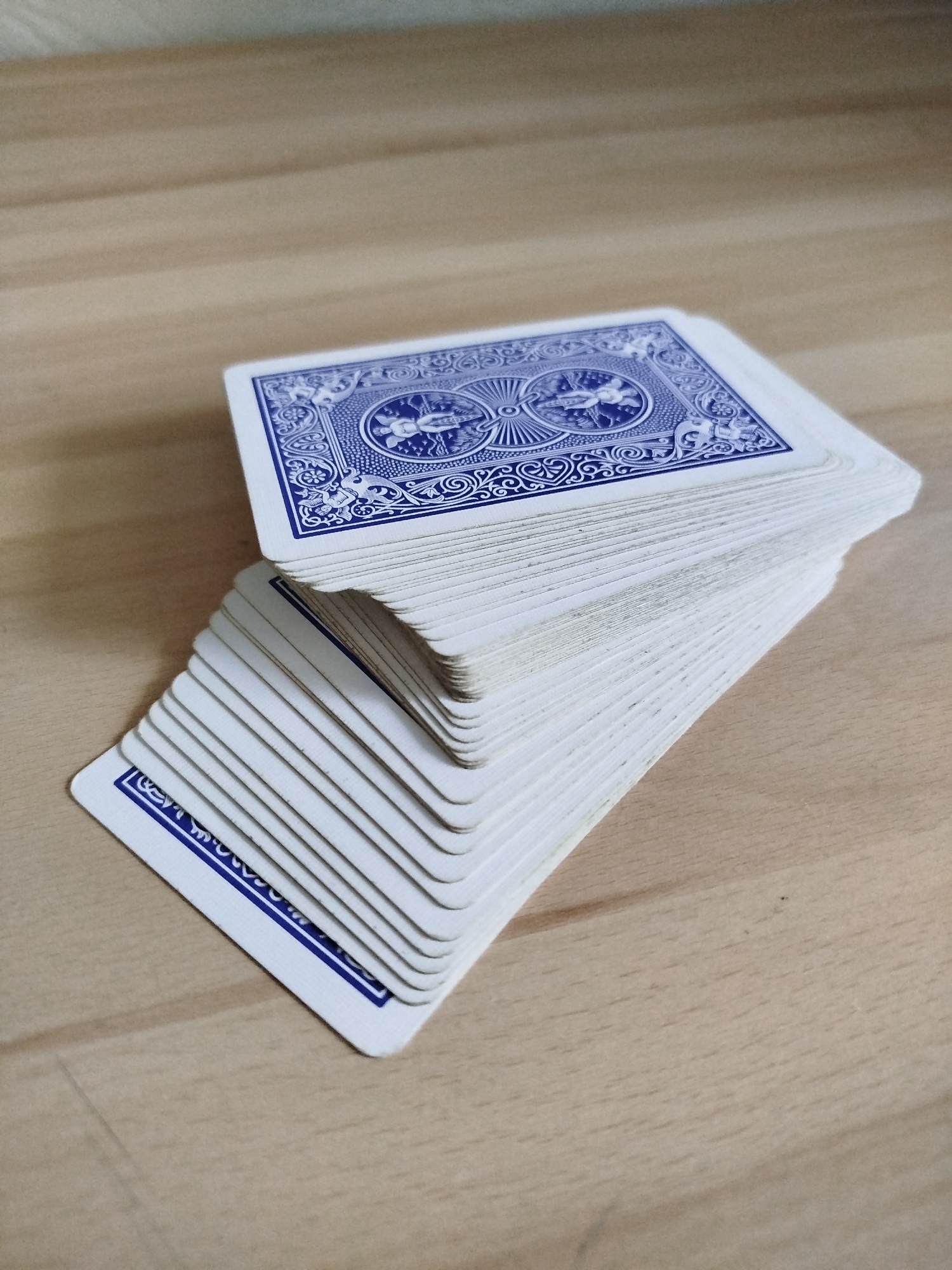 A deck of blue-backed Bicycle brand playing cards rests on a light brown, possibly wooden, surface. The deck is slightly askew, with the bottom portion fanned out. The back design of the top card is visible, featuring a blue pattern with cherubs and a circular design. The edges of the cards appear slightly worn. The scene suggests an indoor environment, likely a table or flat surface in an unknown location. Varna, Bulgaria is not evident in the image.
