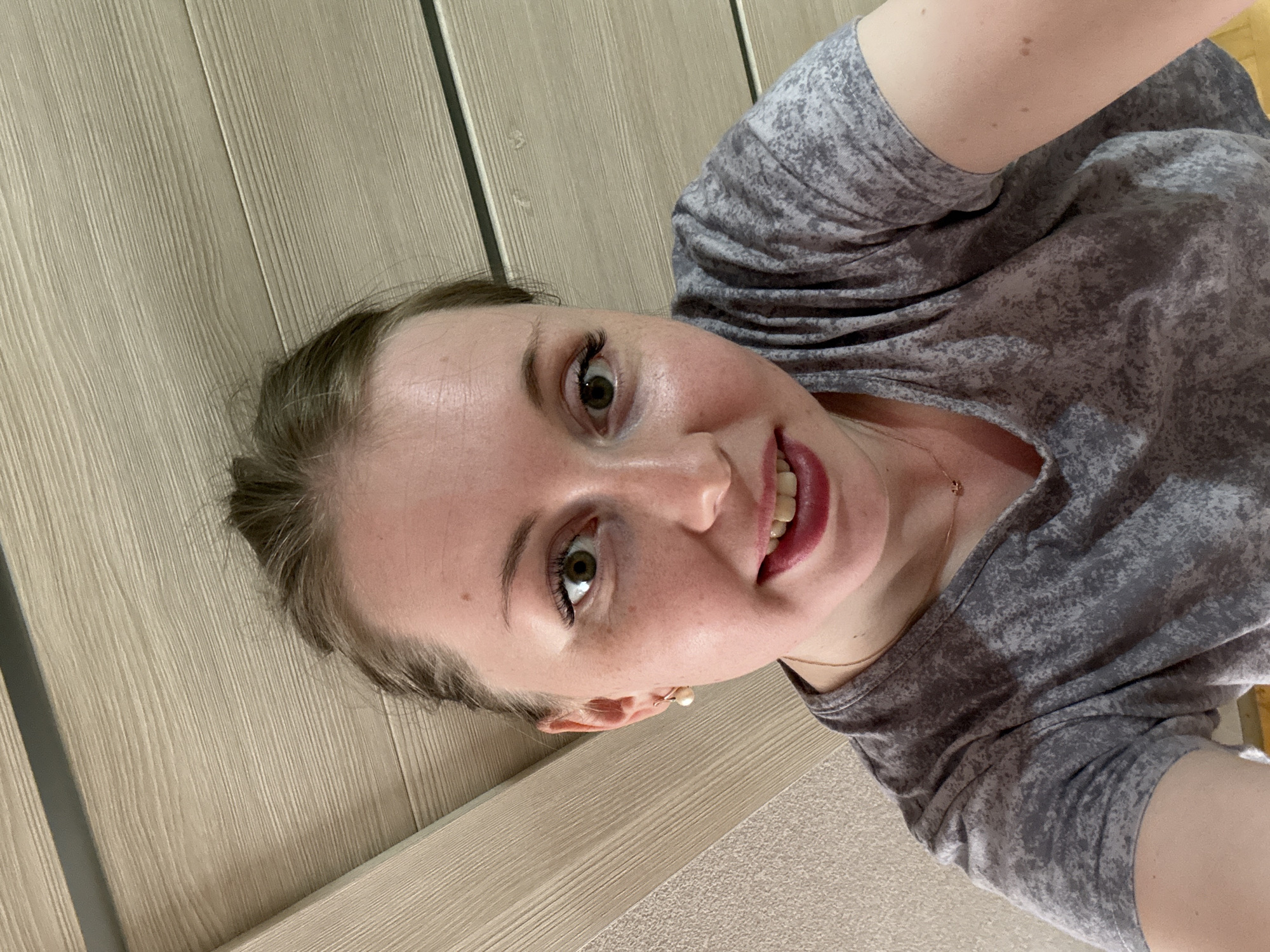 A woman is positioned in front of a beige, wood-paneled wall. She wears a gray patterned t-shirt and a pearl earring. She looks directly at the camera, smiling. The location is Unknown, Unknown.