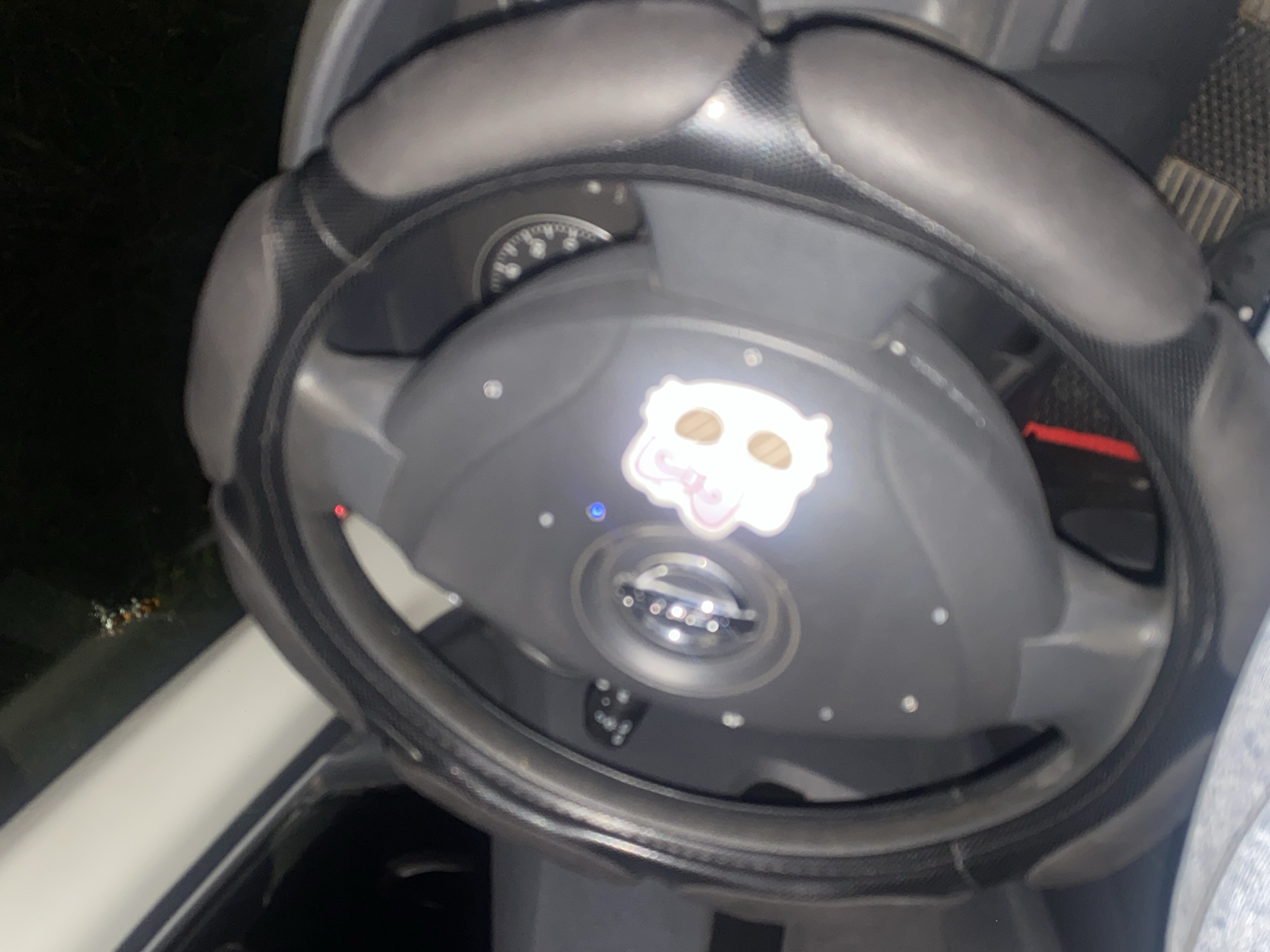 The image shows a close-up view of a car's steering wheel with a protective cover. A sticker with a cartoonish white cat face is affixed to the center of the steering wheel. The background is dark and blurry, suggesting the photo was taken at night or in low-light conditions. Some part of the car's dashboard and interior are visible. The location is specified as Uchaly, Russia, but there are no visual cues in the image to confirm this location.