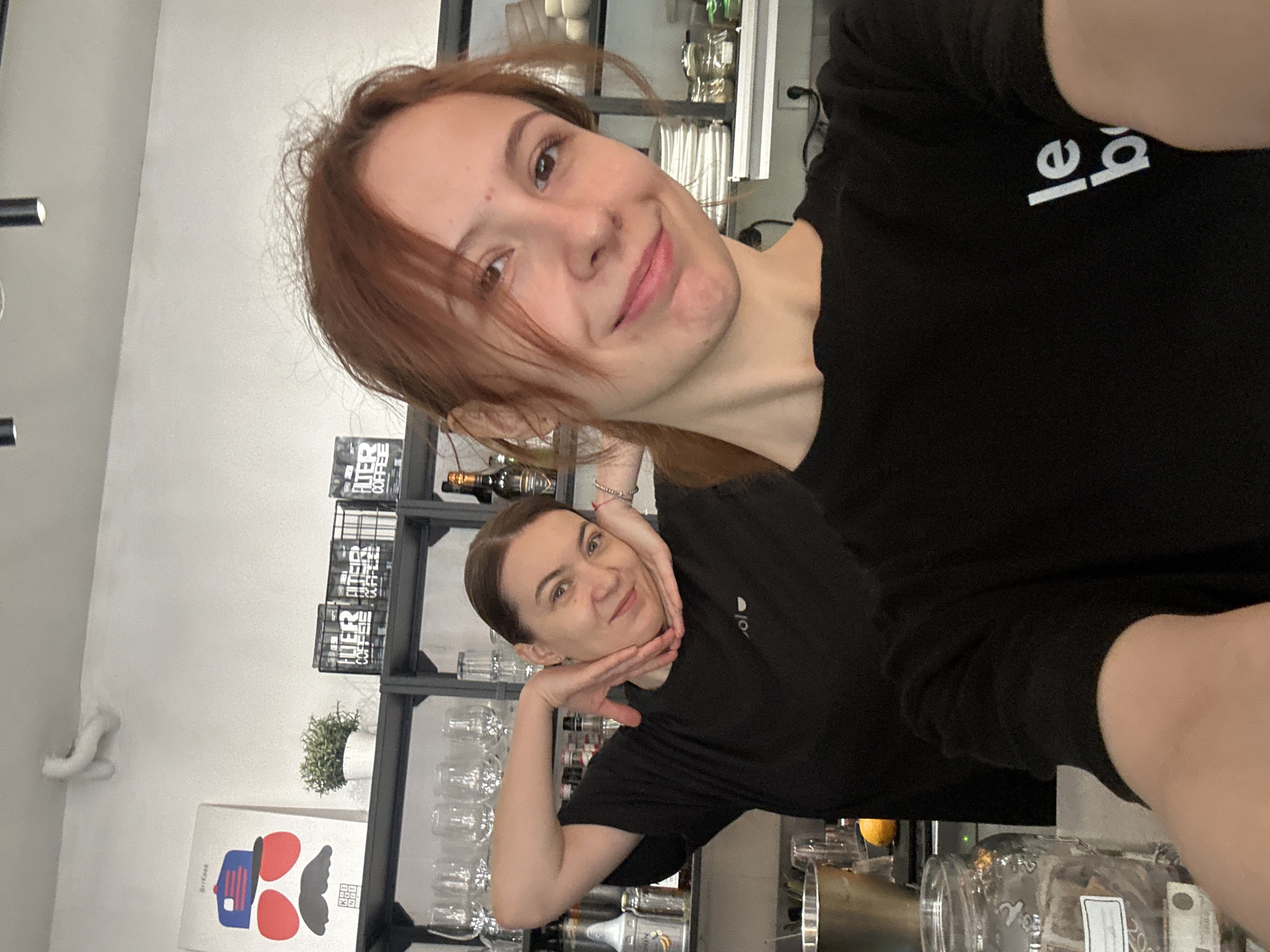 Two women are posing for a selfie in what appears to be a café or restaurant. The woman in the foreground has reddish-brown hair and is wearing a black t-shirt with white lettering. The woman behind her has her hair pulled back and is also wearing a black t-shirt. She has her hands placed on either side of her face.
Behind them is a metal shelf with glasses, bottles, and decorative items, including a small potted plant and containers labelled "FILTER COFFEE". A framed graphic print is on the wall to the left.
Given the "Keep Kafi" print, this photograph was likely taken at a café in Novi Sad, Serbia.