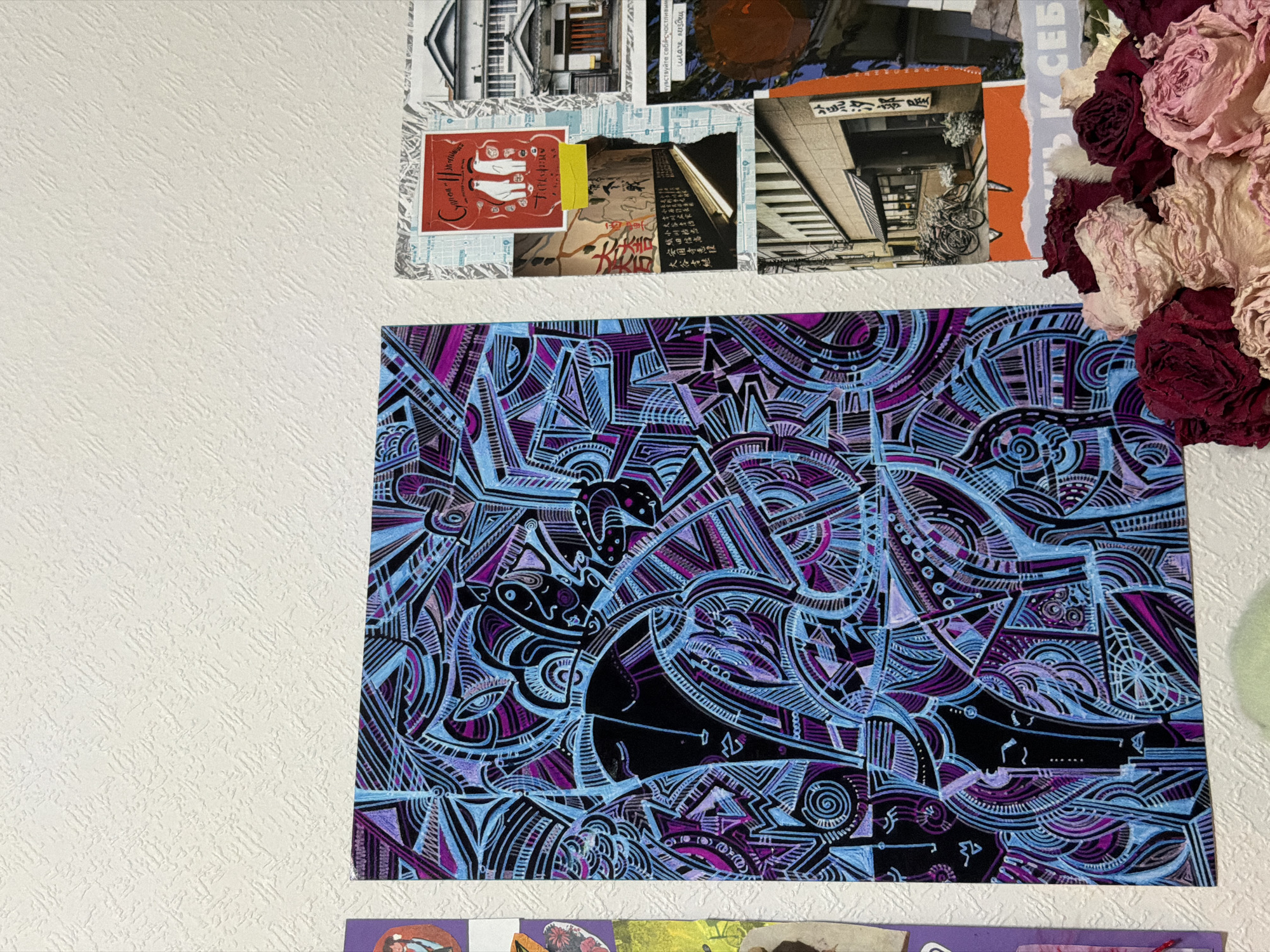 This is a close-up, eye-level, indoor photograph of a collage of artwork and photographs attached to a textured white wall. The dominant feature is a large, rectangular piece of artwork on the left, rendered in black, purple, and light blue with intricate abstract patterns and stylized facial features. To the right of this artwork, a collection of smaller images is arranged. These include a red postcard with two white figures, a photograph of a Japanese street scene with buildings and bicycles, and other torn pieces of paper with text and images. In the foreground, a cluster of dried roses in shades of pink and deep red is visible. The overall impression is of a creative display or mood board. There is no indication of the specific location within Moscow, Russia, or any people or actions occurring in the scene.