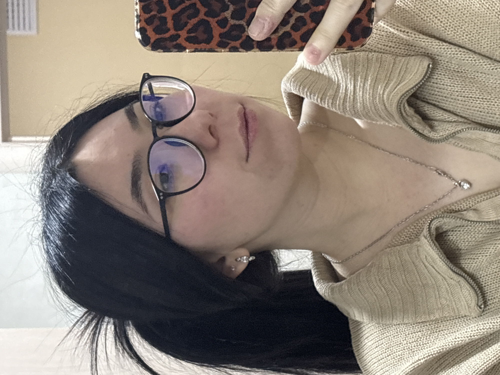 This close-up selfie, rotated 90 degrees clockwise, captures a young person, likely female, looking upwards towards the camera. Her long, dark hair is visible, falling to the side. She wears black-framed, round glasses that reflect light sources, possibly from a screen or window. Her lips are slightly parted, conveying a neutral expression.
She is dressed in a light beige or tan knitted sweater or cardigan, featuring a ribbed texture on the collar and a zipper running along the opening. A silver-colored chain necklace with a small, sparkling pendant adorns her neck. A small stud or hoop earring is visible in her ear. Part of her hand, holding a smartphone with a distinctive leopard print case, is visible at the top of the frame, indicating she is taking the photo herself.
The background is plain and light-colored, suggesting an indoor setting with soft, ambient lighting. No specific time of day or weather conditions can be discerned. The provided context indicates the location of this image is Izhevsk, Russia. There is no visible text in the media file.