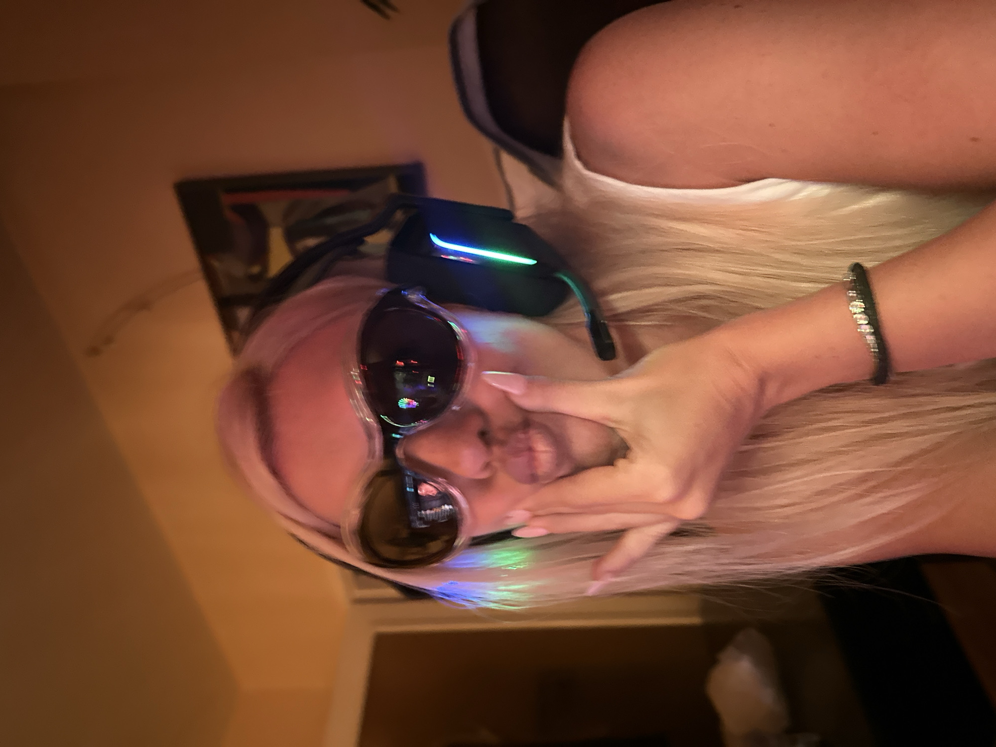In a close-up, medium-shot image, a person with blonde hair and light skin is depicted. They are wearing large, futuristic-style sunglasses with dark lenses and a pinkish frame. A black headset with illuminated blue and green lights is on their head, and a microphone extends from the headset. The person has their left hand placed on their cheek, with their lips puckered in a kissing gesture. They are wearing a white tank top and a thin, dark bracelet with a few rhinestones on their left wrist. The background is dimly lit, showing an orange-toned wall with a framed picture or artwork on it. The lighting suggests an indoor environment, possibly a room with warm ambient light. The image is not taken in Pazardzhik, Bulgaria.