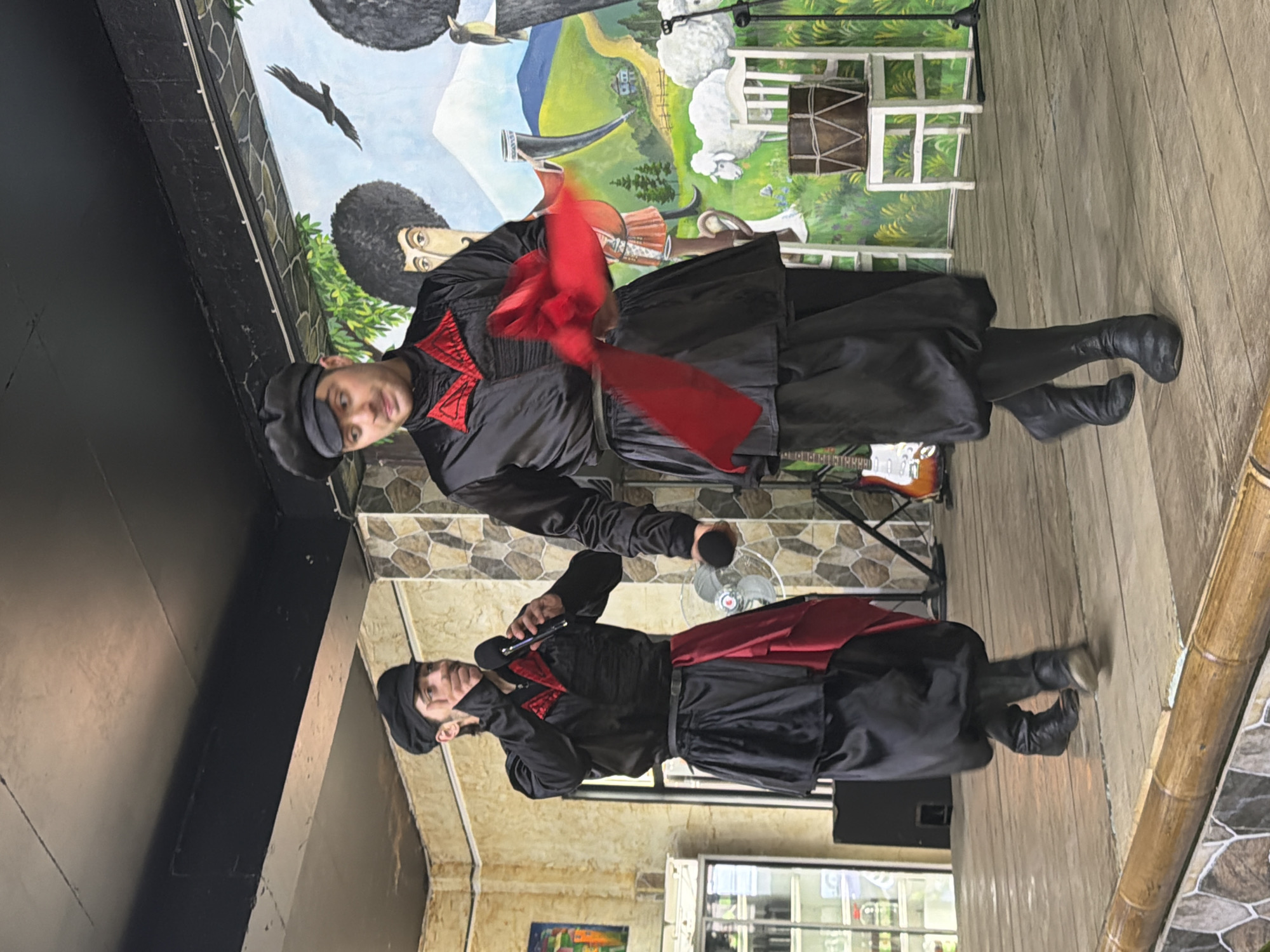 In Kveda Chkhutuneti, Georgia, two individuals are performing on a wooden stage in front of a mural depicting a rural Georgian landscape. The person on the left, wearing a black traditional outfit with red accents and a black cap, is holding a microphone and speaking. The person on the right, similarly dressed in black with red trim and a black cap, is holding a flowing red fabric, possibly for a dance. The mural behind them features stylized figures, sheep, a bird in flight, and a horn. To the left of the stage, a black speaker is visible, and to the right, a white chair with a drum on it and a portion of an electric guitar are present. The lighting suggests an indoor setting, possibly a cultural center or restaurant.