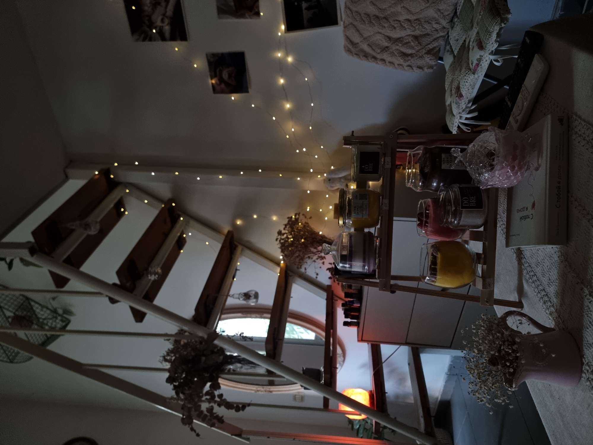 The image shows an interior view, appearing to be within a residence. A staircase with white banisters is visible on the left, leading upwards. A window with a semi-circular arch is present. Decorative lights are strung across the ceiling. A wooden shelf unit is visible, displaying multiple jars and decorative objects. A book is placed on a surface near the shelf, and a small vase with dried flowers sits beside it. The image is not geographically indicative of Tel-Aviv.