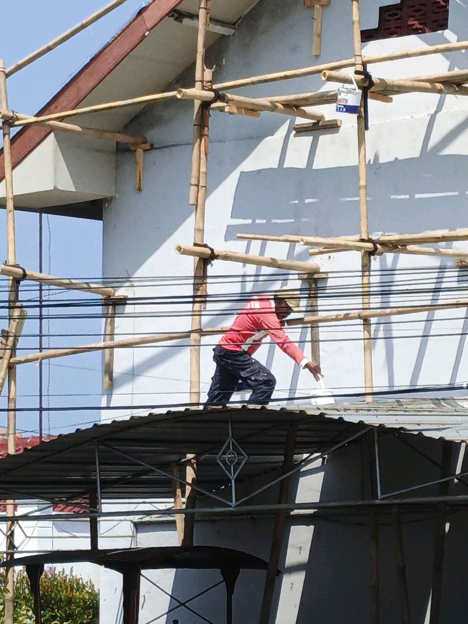 A construction worker in a red long-sleeved shirt and dark pants is standing on scaffolding, reaching down to paint a surface. The scaffolding is made of bamboo poles tied together with rope. There are electrical wires strung across the scene in front of the scaffolding. The worker is near the top of a sloped, corrugated metal roof structure. Behind the scaffolding, a white wall of a building is visible, with shadows cast by the scaffolding. A small white can with blue lettering is hanging from one of the bamboo poles. The sky is clear and blue. The location is stated as Desa Tegalreja, Kecamatan Cilacap Selatan, Indonesia.