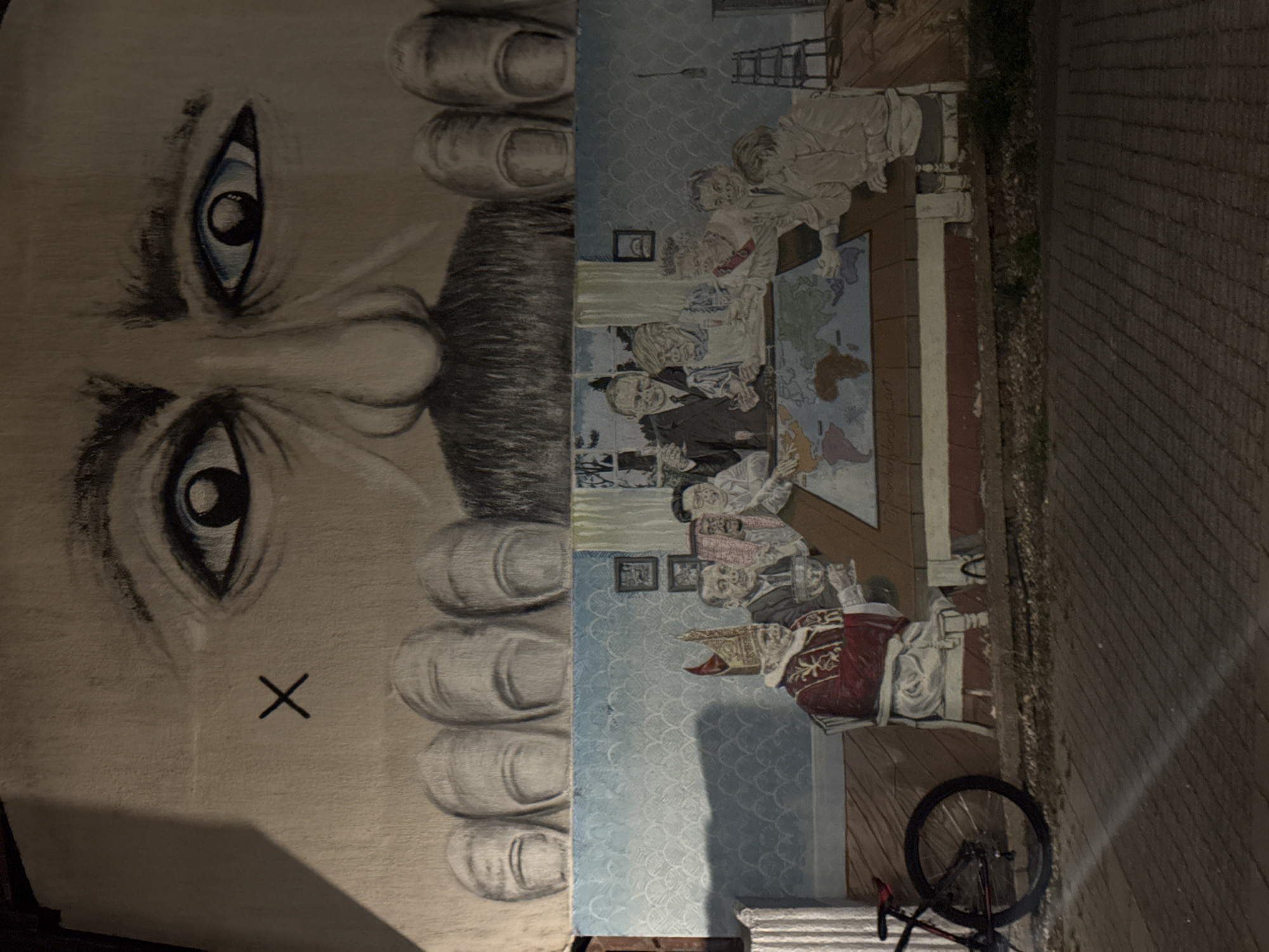 The image shows a mural on a building at night. The mural depicts a large face with prominent eyes and a mustache at the top, with several fingers visible below. The lower portion of the mural features a scene with several people gathered around a table with a world map on it. One person is dressed in religious vestments, resembling a pope, and is holding a chalice. Other figures appear to be leaders or dignitaries, engaged in discussion or observation of the map. The backdrop of this lower scene includes patterned wallpaper and a window showing a treeline. To the left of the mural, a bicycle with a red frame is parked. The ground in front of the mural is paved with bricks and appears to be a sidewalk or street. The mural is located in the Zvezdara Urban Municipality, Serbia.