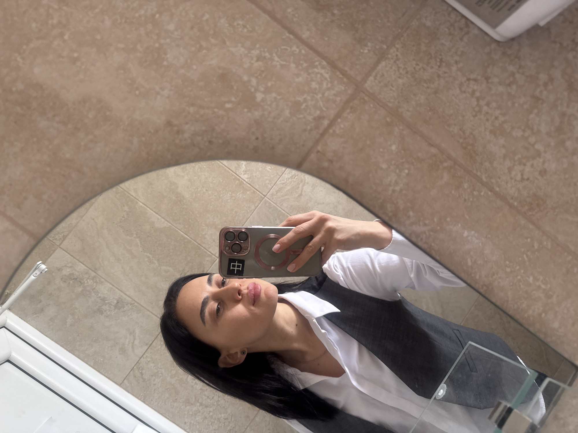 The image is a selfie taken in a bathroom mirror. A woman with dark hair is holding a phone with a rose-gold case and a Chinese character sticker in the upper left corner. She is wearing a white shirt and a dark gray vest. The bathroom has beige tile walls and a white door frame. The location cannot be determined from the image.