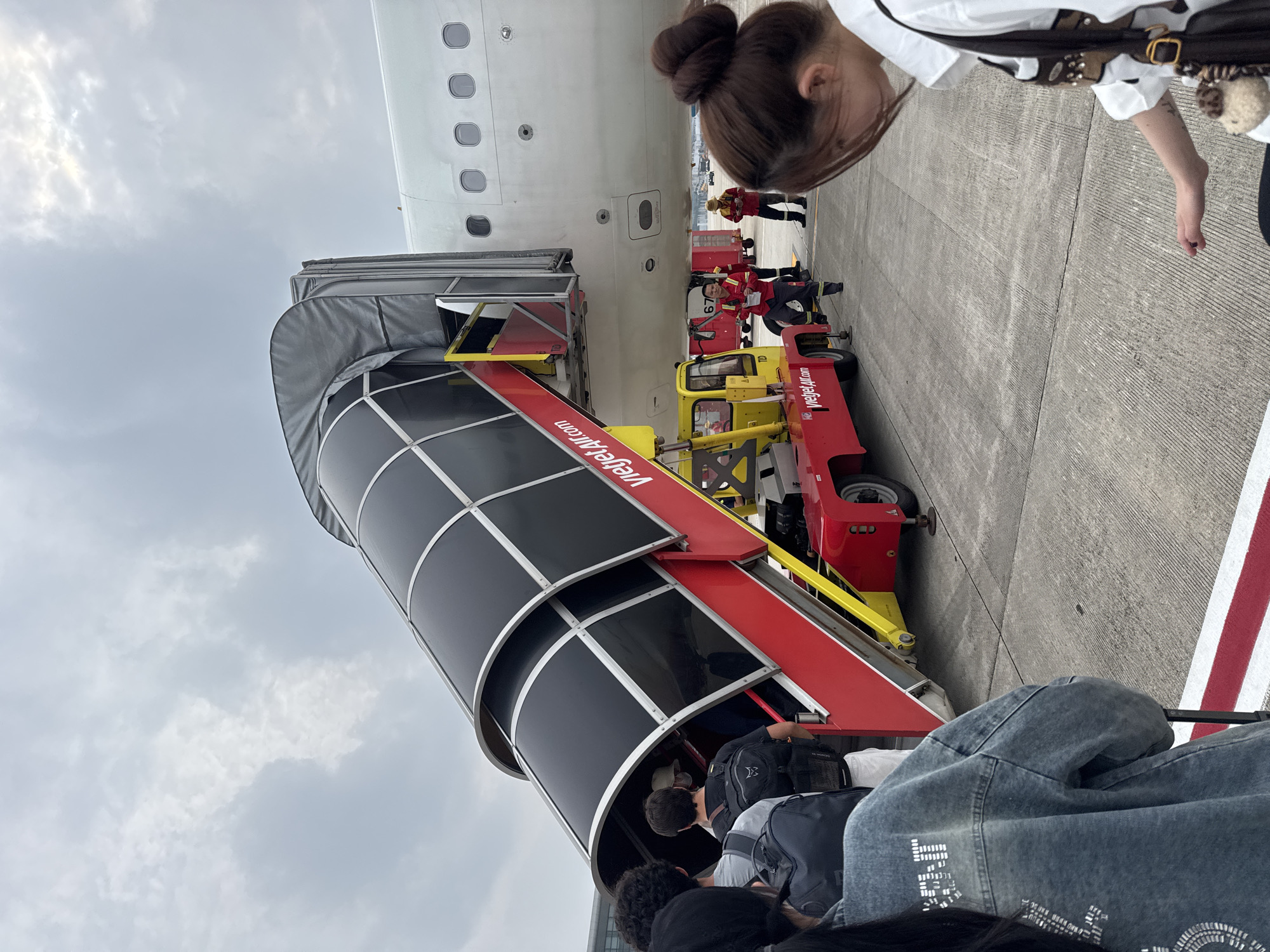 An airplane boarding ramp, branded with "Vietjet Air," is attached to a white airplane. People are seen boarding the plane via the ramp. A yellow and red vehicle is positioned beneath the ramp, also bearing the "Vietjet Air" logo. Airport personnel in red uniforms are visible near the vehicle and the ramp. The scene takes place on an airport tarmac, under an overcast sky. The location is identified as the Noi Bai International Airport in Hà Nội, Vietnam.