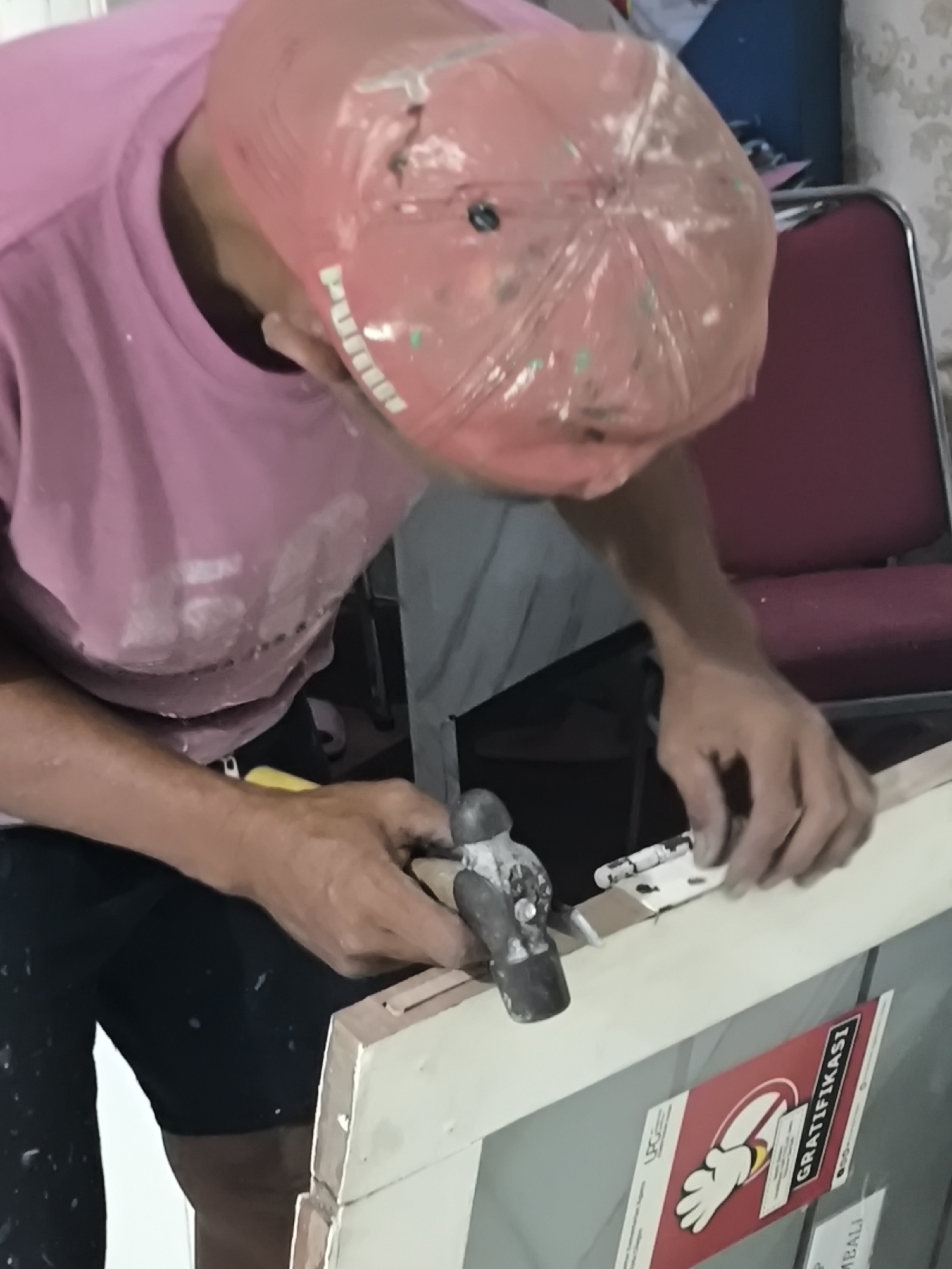 In a room in Cilegon, Indonesia, a person wearing a pink t-shirt and a red cap is working on a window frame. The person is holding a hammer in their right hand and appears to be attaching a hinge to the frame. The window frame is made of wood and is light brown in color. There is a sign with the word "GRATIFIKASI" on it, which is Indonesian for "gratification" or "reward," visible on the window. The room appears to be indoors, with a chair and a patterned wallpaper visible in the background. The person's hands and clothing are stained with white paint, suggesting they are engaged in a painting or construction activity.