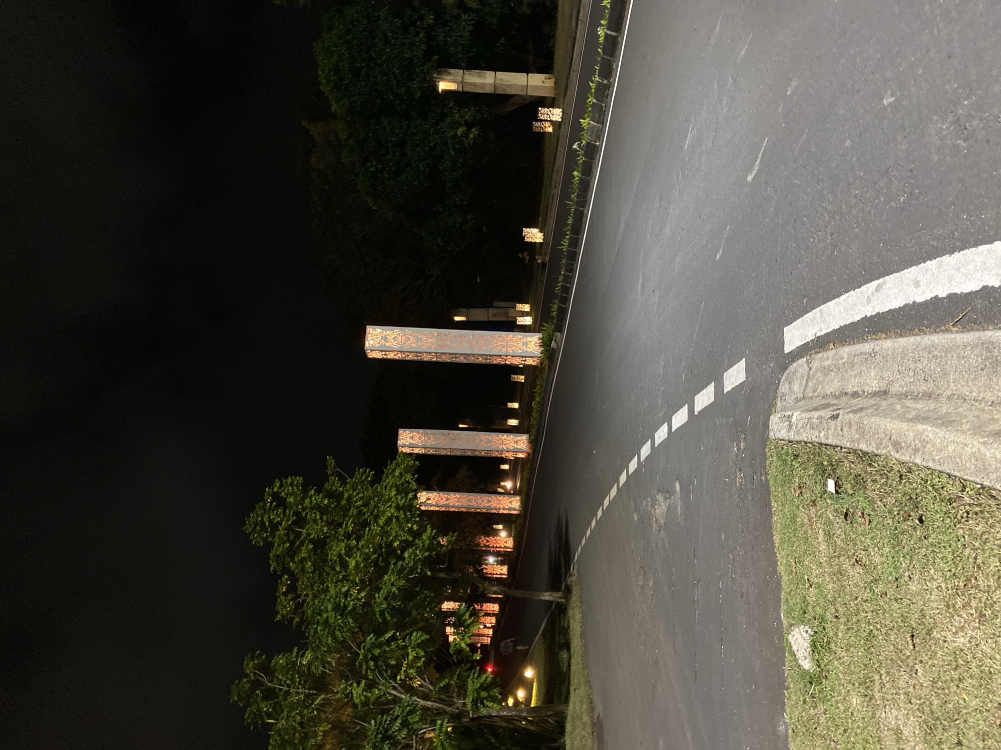 The image depicts a nighttime scene on a paved road in Kuta Selatan, Indonesia. Tall, illuminated pillars with intricate patterns line the right side of the road, serving as decorative lighting. The road features white dashed lines, indicating traffic lanes. To the left, trees and grass are visible, with scattered ground lighting adding to the ambiance. A single person is observed walking on the left side of the road, near the illuminated pillars. The overall environment suggests an outdoor public space, likely a road leading to a venue or an area with ornamental landscaping. The sky is dark, indicating nighttime conditions.