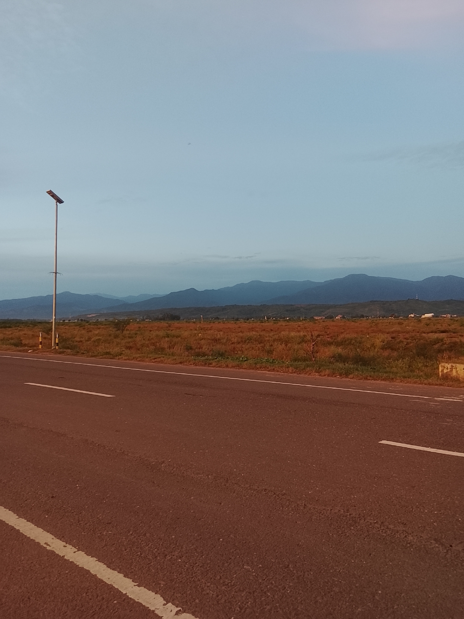 An outdoor, medium-shot, eye-level, vertical, portrait-orientation photograph captures a rural landscape at twilight. In the foreground, a dark asphalt road with white lane markings stretches diagonally from the bottom left to the upper right. To the left of the road, a tall, silver lamppost with a solar panel stands beside a yellow and black striped pole. Beyond the road, a wide expanse of dry, golden-brown grass and sparse vegetation extends towards a distant mountain range. The mountains, rendered in muted blue and gray tones, form a hazy silhouette against a pale, gradient sky that transitions from a light pinkish hue at the top to a pale blue at the horizon. A few wispy clouds are visible in the upper sky, and a very small, faint object, possibly an airplane or bird, is visible in the upper left quadrant of the sky. The scene appears to be located in the vicinity of Dayah Kruet, Indonesia, given the rural setting and mountainous backdrop.