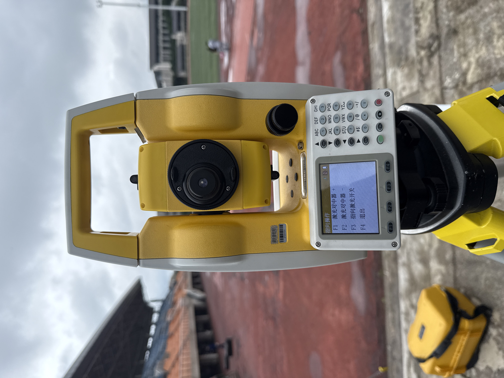 A yellow and white total station, a surveying instrument, is mounted on a tripod. The total station has a screen displaying Chinese text and a keypad. In the background, there is a stadium with orange seats and a cloudy sky. A yellow case, presumably for the instrument, is visible on the ground to the left. The scene appears to be set up for surveying work within the stadium grounds in Jiangjin, China.