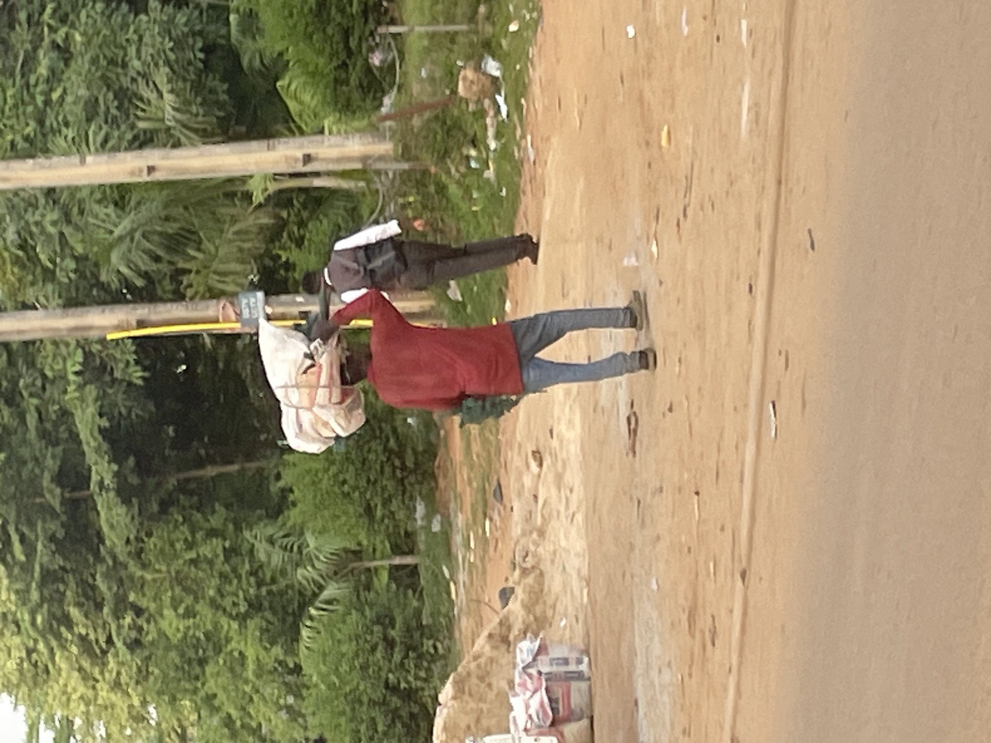 In a dusty urban environment in Abuja, Nigeria, two individuals are walking. In the foreground, a person with dark skin wears a red long-sleeved shirt and blue jeans, carrying a large bundle of wrapped items balanced on their head. They are walking away from the viewer and towards the right of the frame. A green bag is slung over their shoulder. In the midground, a second person, also with dark skin, walks away from the viewer, wearing a white shirt, a dark vest, and dark pants. They have a dark backpack on their back. Behind the individuals, the scene is characterized by lush green trees and vegetation. There are utility poles with some signage, and piles of dirt and bagged materials, possibly cement, are visible on the left. The ground is a mix of sand and dirt. Litter is scattered across the ground, suggesting a less-than-pristine urban setting.