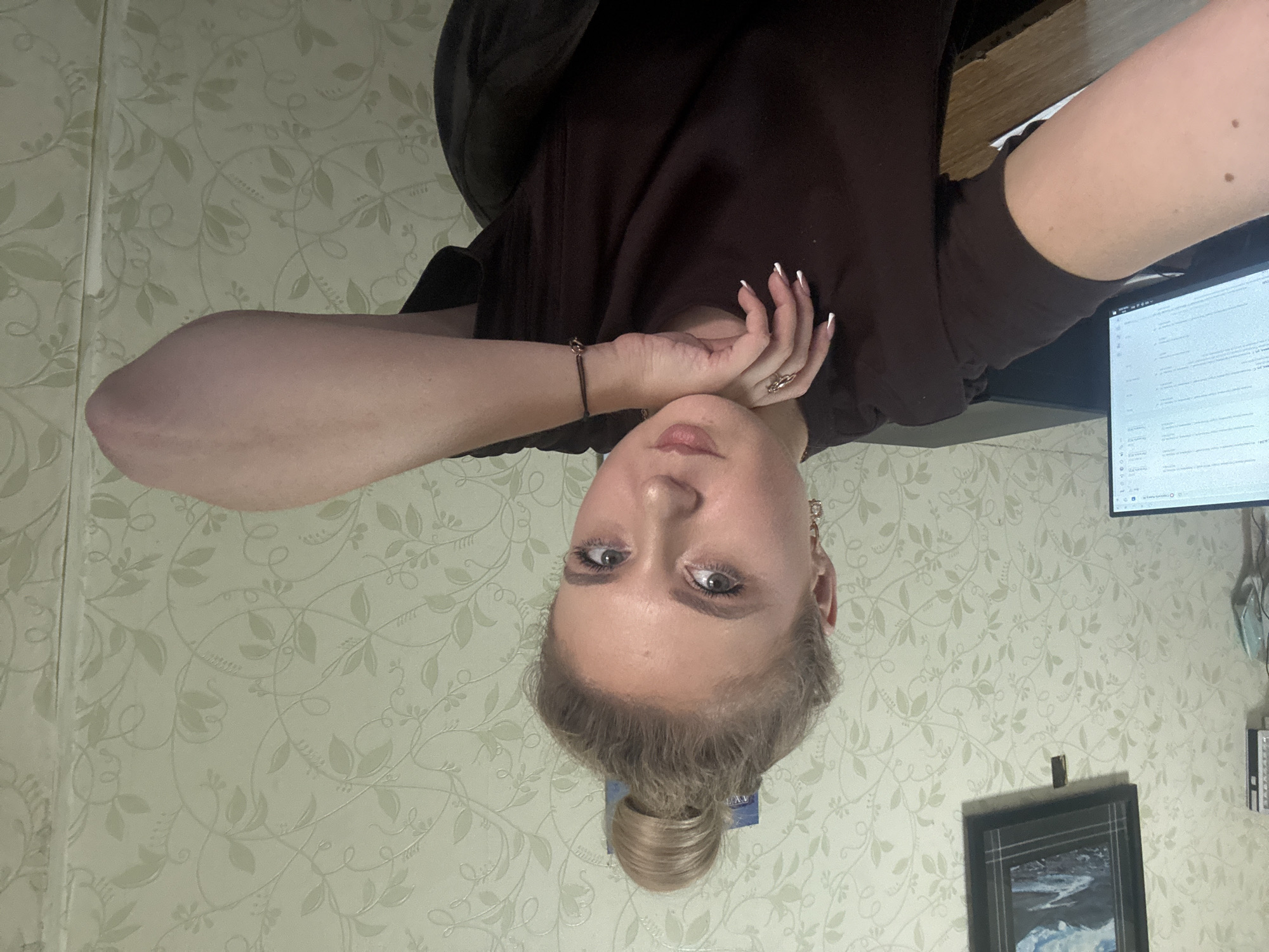 A young woman with blonde hair styled in a bun is leaning on her elbow, resting her chin on her hand. She is wearing a dark brown t-shirt and a ring on her finger. A bracelet is visible on her wrist. She has long fingernails painted with white tips. The background consists of patterned wallpaper and a computer monitor is visible to her left. A framed picture hangs on the wall above and to the left of the woman. The scene appears to be indoors, possibly an office or home environment.