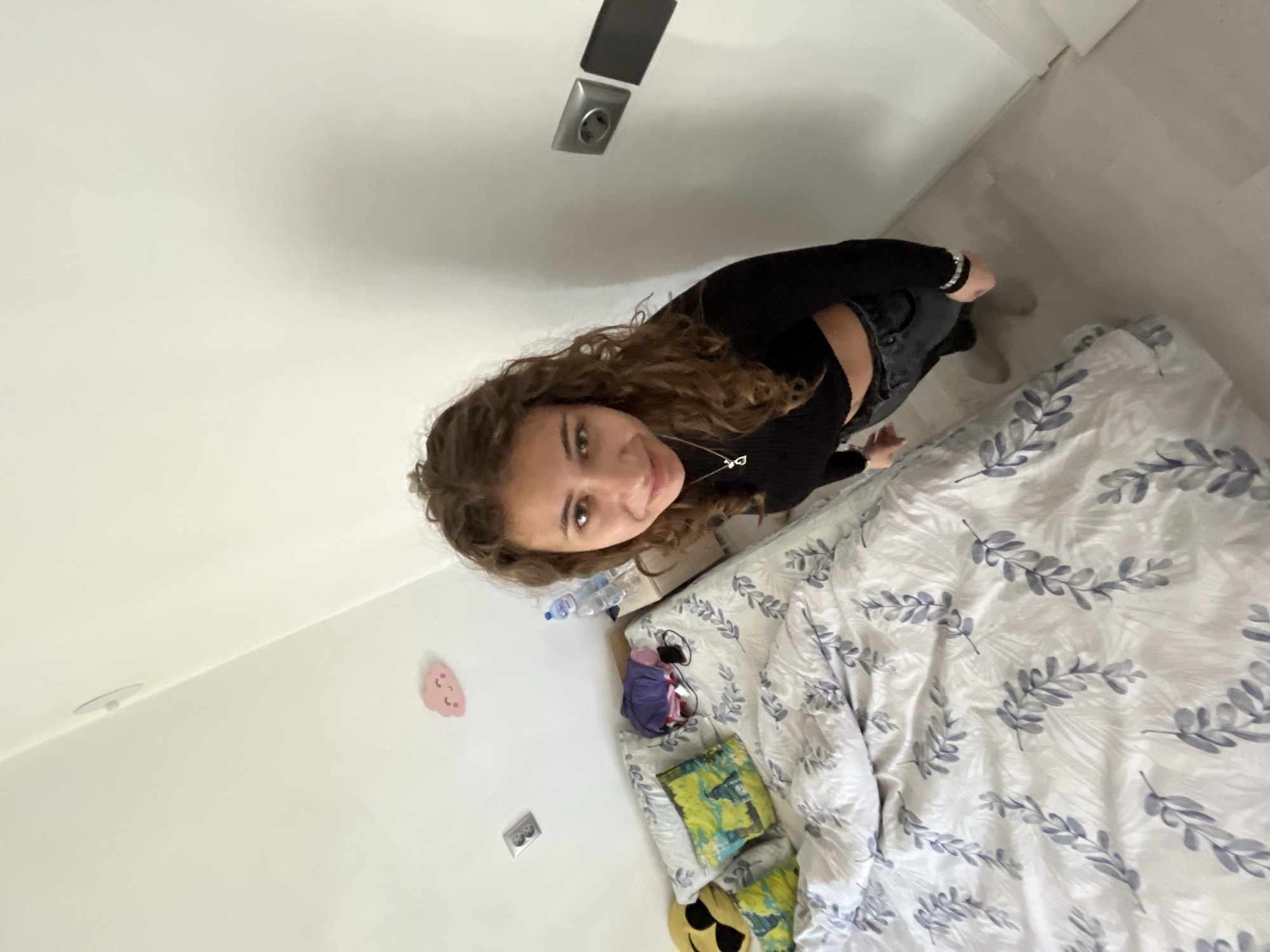 A young woman with curly brown hair is positioned in a room, looking directly at the camera. She is wearing a black, long-sleeved crop top and dark jeans. Behind her is a bed covered in a white duvet with a pattern of blue-grey leaves. The room has white walls with a pink wall sticker and a power outlet. The location is in a room, likely a bedroom, in Karavelovo, Bulgaria.