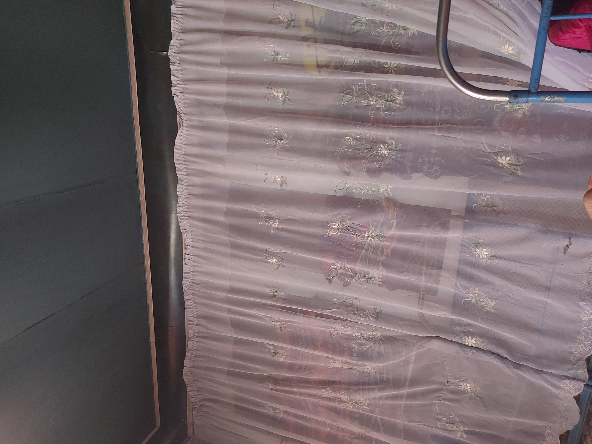 The image depicts an interior scene with a dark teal ceiling composed of wooden planks. A thin wooden trim separates the ceiling from the wall, which is covered by a sheer, light pink curtain. The curtain is adorned with delicate white floral embroidery. The curtain hangs from a rod and is partially drawn, revealing a blurred background that suggests a window or another room. To the right of the curtain, a portion of a blue metal bed frame is visible. The lighting appears to be artificial and ambient, casting soft shadows. There are no people or clear actions taking place in the frame. The scene is domestic and appears to be in a residential setting, possibly a bedroom. The location is identified as Karkidon, Uzbekistan.