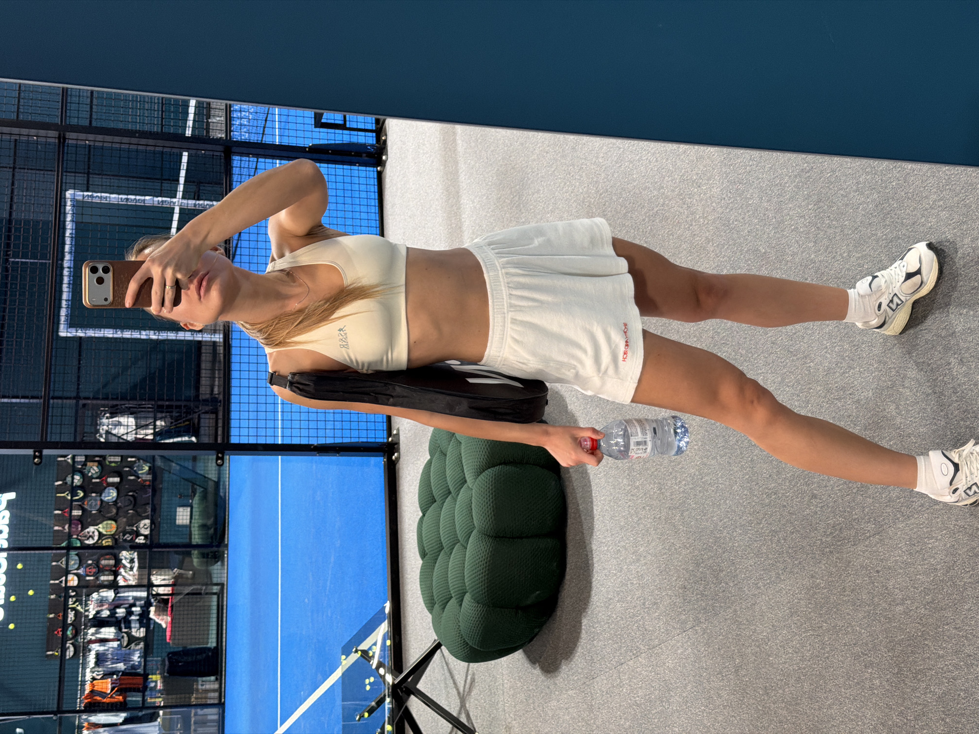 A woman is taking a selfie in what appears to be an indoor paddle tennis facility. She is wearing a light-colored sports bra and tennis skirt, white socks, and athletic shoes. She is holding a cell phone in her right hand and a water bottle in her left. A black tennis bag is positioned beside her. Behind her, paddle tennis courts are visible through a glass or transparent barrier. To the left, a display case of tennis racquets and apparel is present. A dark green, tufted ottoman-like seat is located near the woman. The floor is a speckled grey surface. Based on the branding visible in the reflection, the location is in Moscow, Russia.