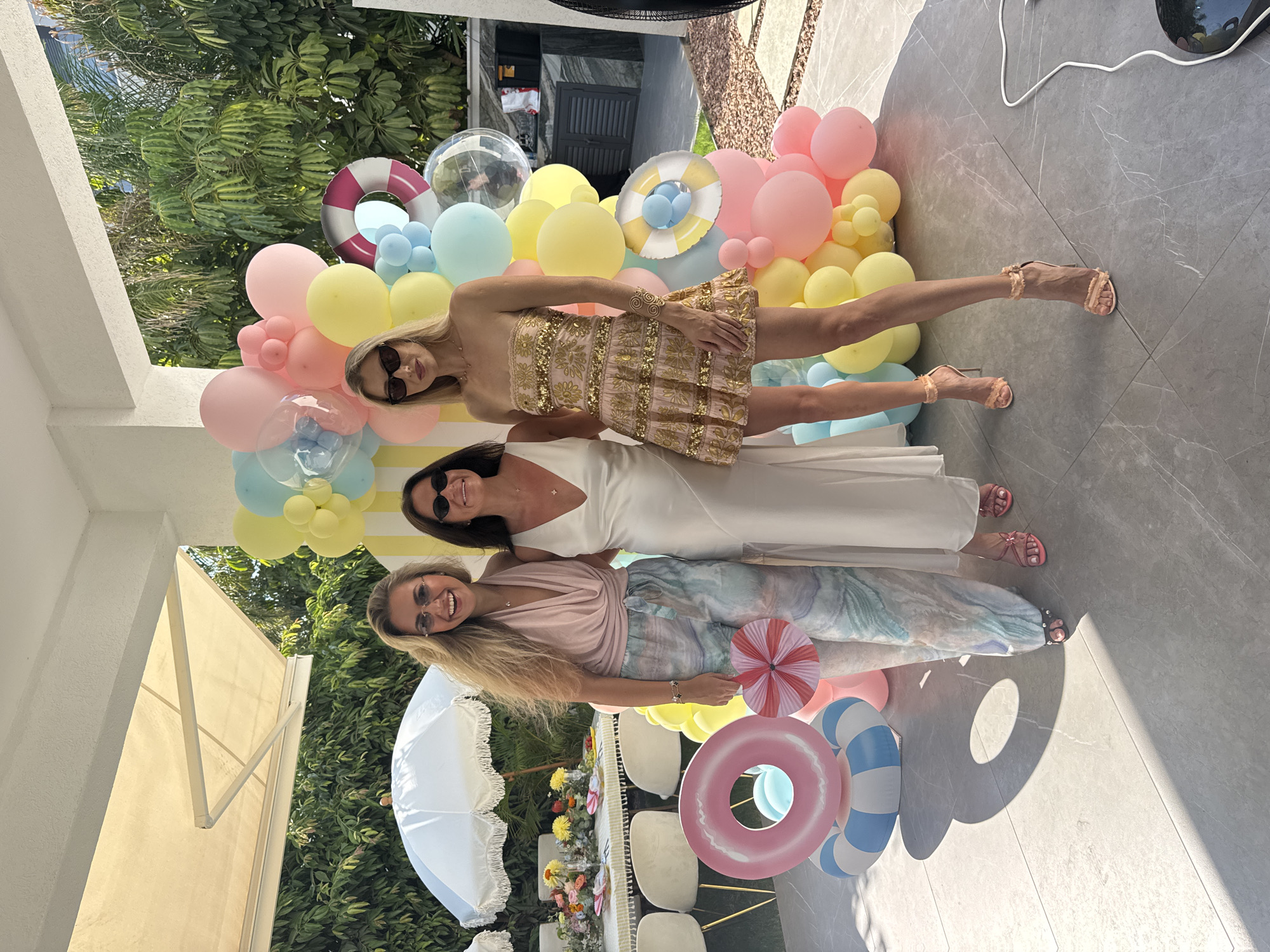 Three women stand in front of a balloon arch and a table set for a meal outdoors. The woman on the left wears a pale pink halter top and patterned wide-leg pants. She holds a pink striped paper fan. The woman in the middle wears a long, white V-neck dress. The woman on the right wears a strapless pink mini-dress adorned with gold sequins and embroidery. She has a gold cuff on her left wrist. The background shows a covered patio area with a table set with floral arrangements and white chairs. Behind the women is a large balloon display in pastel colors, including pink, yellow, and blue. There are also inflatable pool rings at the base of the balloon arch. The ground is paved with large gray tiles. The overall scene appears to be a celebration or party.