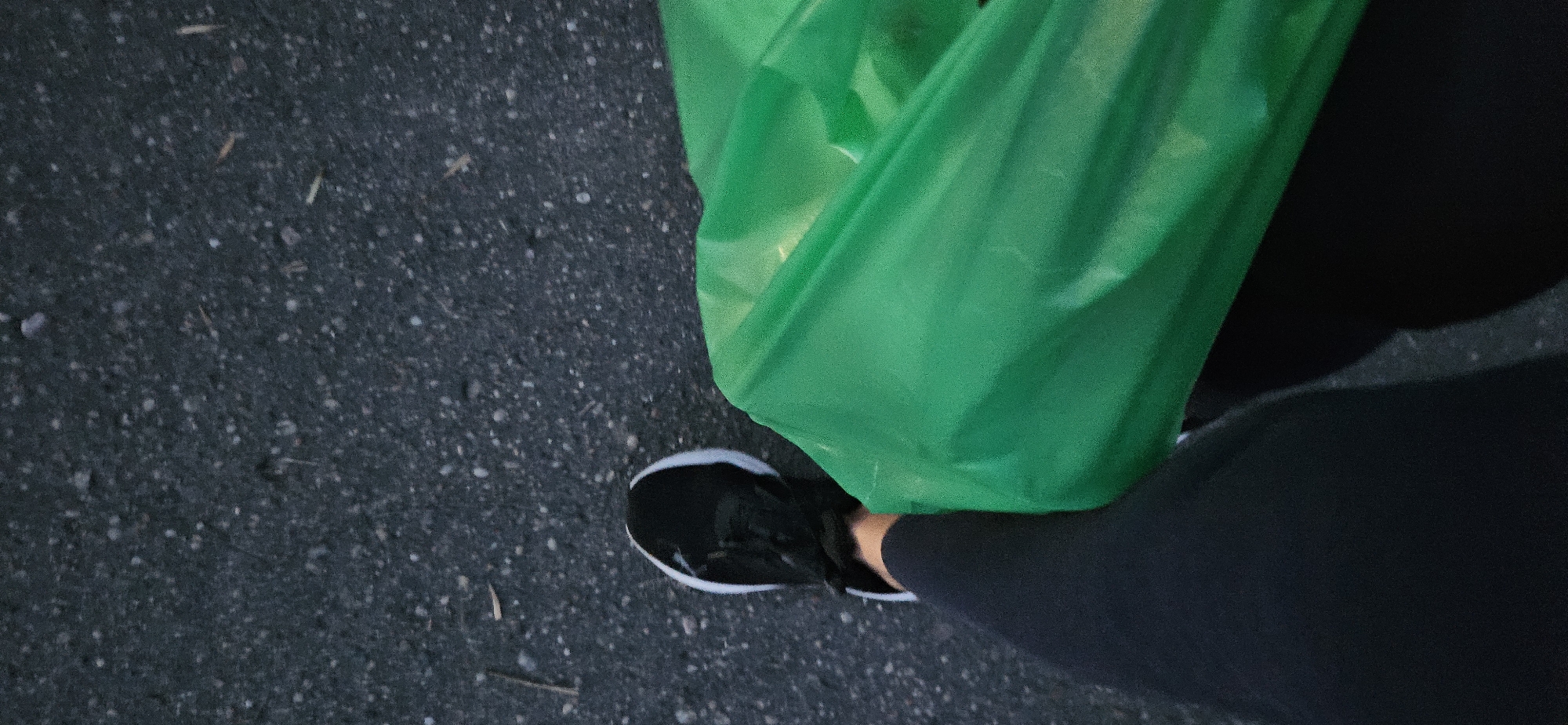 The image is a first-person perspective shot, showing the legs and feet of an individual walking on an asphalt surface. The person is wearing black leggings and black and white athletic shoes. To the right of the person's legs is a bright green bag, possibly made of plastic or fabric, partially obscuring the lower part of the right leg. The asphalt appears dark and textured, with small pebbles and occasional dry grass or debris scattered on its surface. The lighting suggests it is either early morning or late evening. There is no discernible location in the city of Vitsebsk, Belarus, identifiable from the image.