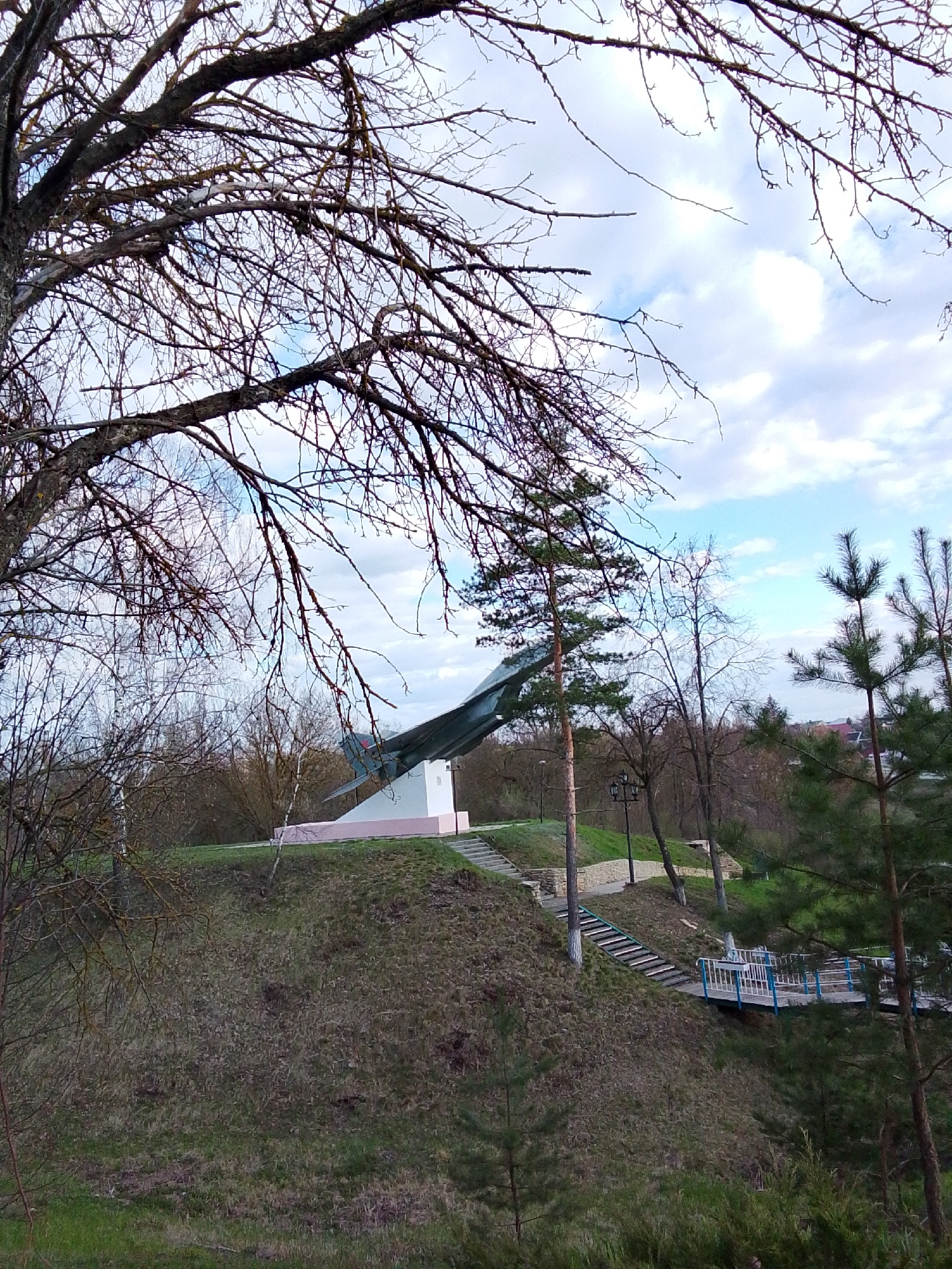 This outdoor photograph captures a commemorative monument set within a park-like landscape in Khlevnoye, Russia. The central subject is a military jet, likely a fighter aircraft, displayed on a pedestal atop a grassy hill. The pedestal has a white upper section and a pink base, with the jet angled upwards as if in flight.
The foreground is framed by the dense, bare branches of large deciduous trees, suggesting late autumn or early spring. Several evergreen trees, including a tall pine, are also visible on the hillside. The ground is covered in a mixture of dry and green grass, indicating a transition season.
To the right of the monument, a set of stone or concrete stairs ascends the hill, bordered by a stone wall. Further down the slope, another pathway or stairs leads to a small footbridge with distinctive blue and white railings. A black lamppost stands along one of the pathways. In the far background, beyond the trees, glimpses of distant buildings or rooftops can be seen under a partly cloudy sky, with patches of blue visible between white clouds.
No people are present in the image, and no specific activity is occurring, conveying a sense of quiet solitude. The lighting indicates it is daytime, possibly morning or late afternoon. There is no visible text in the photograph.