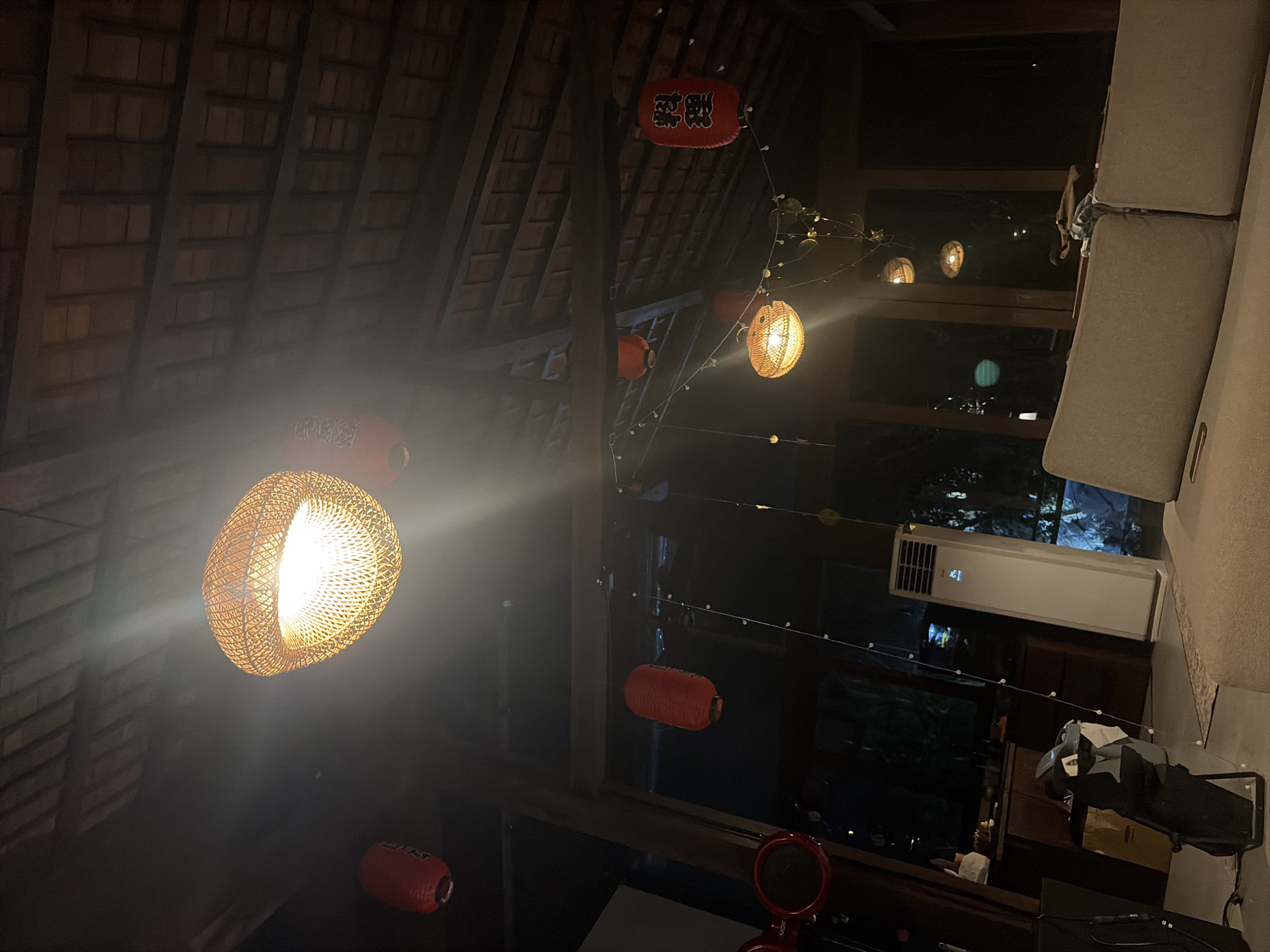 The image depicts an indoor scene with a rustic, dimly lit ambiance. The ceiling, constructed of wooden beams and what appears to be thatched roofing, suggests a traditional or bohemian architectural style. Several decorative lanterns are suspended from the ceiling. One prominent lantern, made of woven material, emits a warm, bright light and is centrally located in the upper portion of the frame. Other smaller, red, cylindrical lanterns are also visible, some with Japanese characters. In the background, there is a tall, white air conditioning unit. The foreground is occupied by a light-colored sofa, with a brown bag resting on its back. The lighting is predominantly from the lanterns, creating a warm glow and casting shadows. The overall impression is one of a relaxed, indoor setting, possibly a living room or lounge area, illuminated by decorative lights. The reference to Kediri, Indonesia, suggests the image was taken in a location within that city.