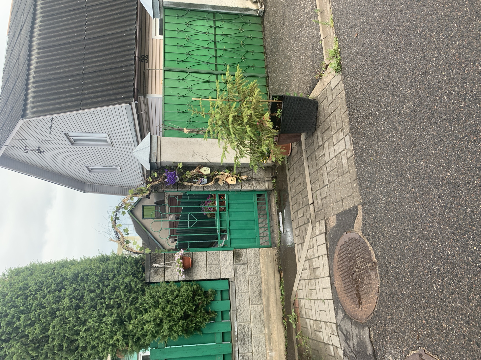 A wide outdoor shot captures a residential property entrance in Minsk, Belarus, on a cloudy day. The foreground shows a wet asphalt road and a paved walkway with a manhole cover. To the right, a green, ornate metal gate and fence border the property. A small evergreen tree in a dark pot stands near the gate. The walkway leads to a green metal gate, which is partially open, revealing a small courtyard beyond. A stone wall and a tall evergreen tree stand to the left of the gate. Behind the gate and tree, a two-story house is visible with light-colored siding and a dark, corrugated roof. The house has a house number "32" visible. The scene is domestic and appears well-maintained, with some potted plants and decorative elements like birdhouses and a woven archway. There are no people or animals visible in the image.