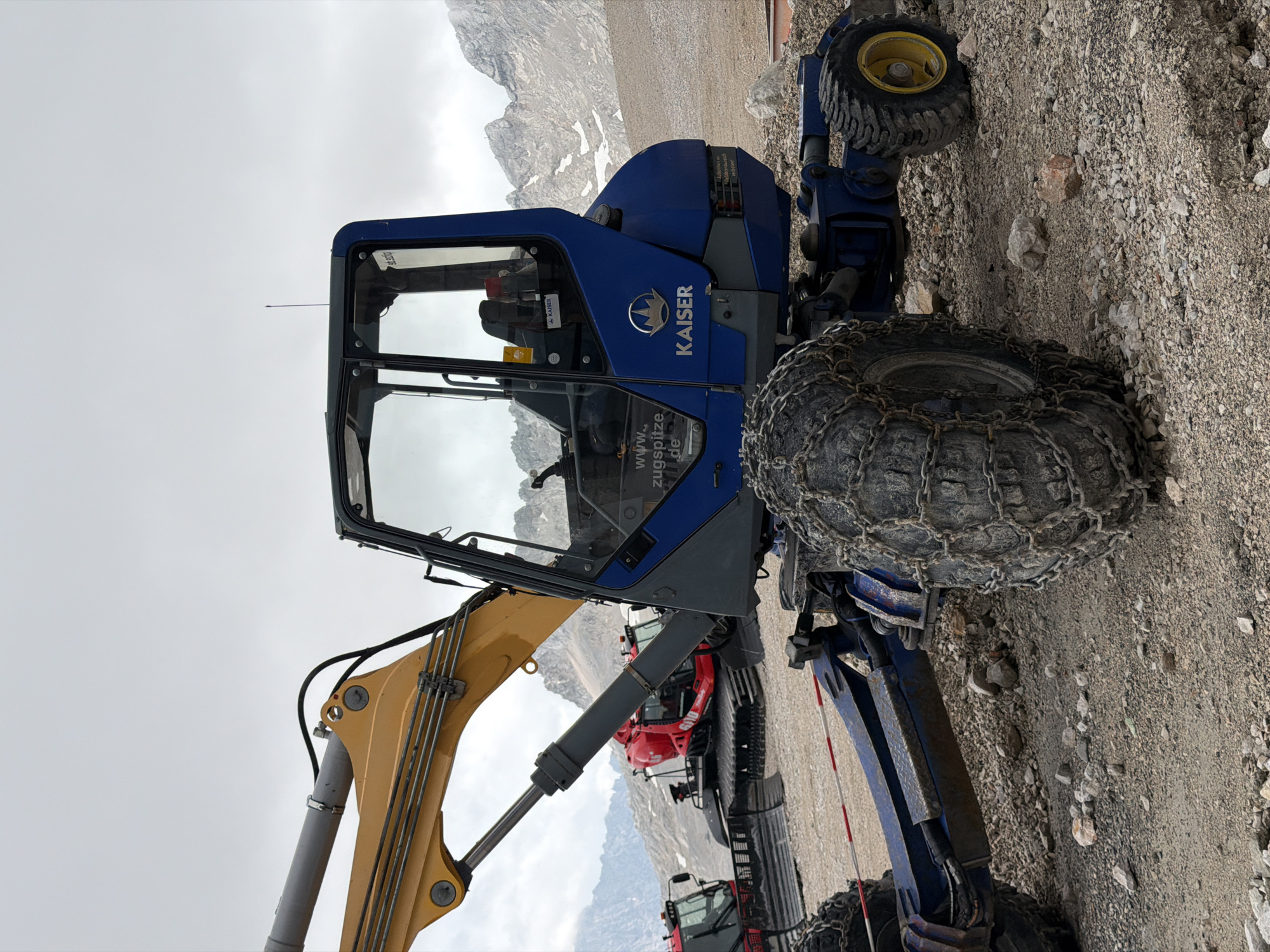 The image shows a blue and yellow excavator with heavy-duty tire chains, parked on a gravelly surface. In the background, two red snowcats are visible, also on the gravel. The scene appears to be in a mountainous area, with rocky slopes and a cloudy sky. The excavator has a logo that reads "KAISER". The overall environment suggests a high-altitude location, possibly a mountain resort or a construction site in a rugged terrain. The presence of snowcats hints at a location that experiences snow, and the excavator is likely used for maintenance or construction work in such conditions. The location in Garmisch-Partenkirchen, Germany, likely refers to the Zugspitze, Germany's highest peak, which is accessible by mountain railways and is a popular tourist destination known for its winter sports and challenging terrain.