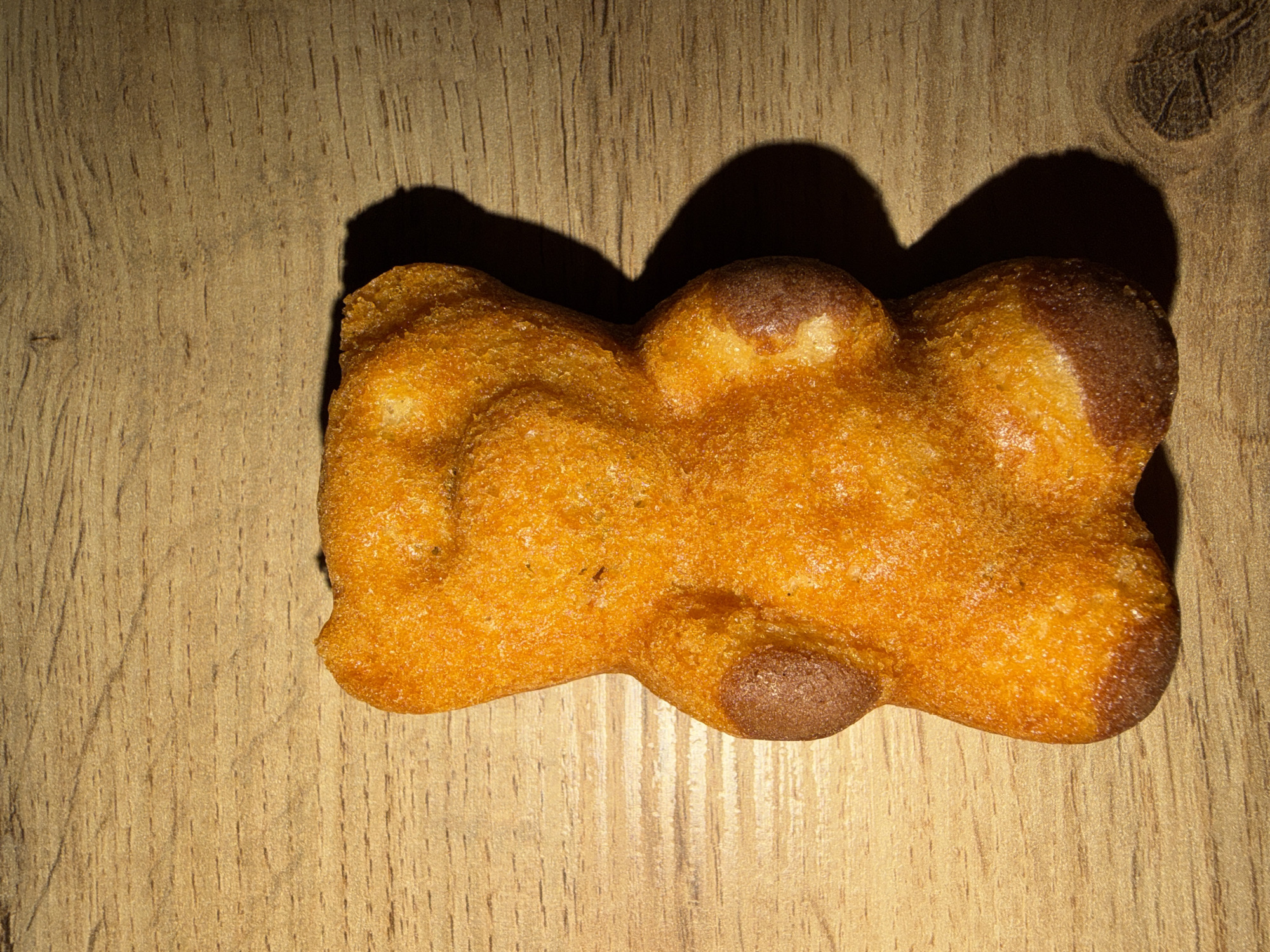 A close-up, still life photograph captures a single, golden-brown, irregularly-shaped object on a light-brown, wood-grain surface. The object appears to be a baked good or snack item, with a slightly uneven texture and darker coloration on its rounded edges. It casts a defined shadow behind it, suggesting a focused light source from above and to the right. The scene is minimalistic, focusing solely on the object and its immediate surroundings, lacking any contextual clues to determine a specific location. It is impossible to determine the location in the city of Plovdiv, Bulgaria from this image alone.