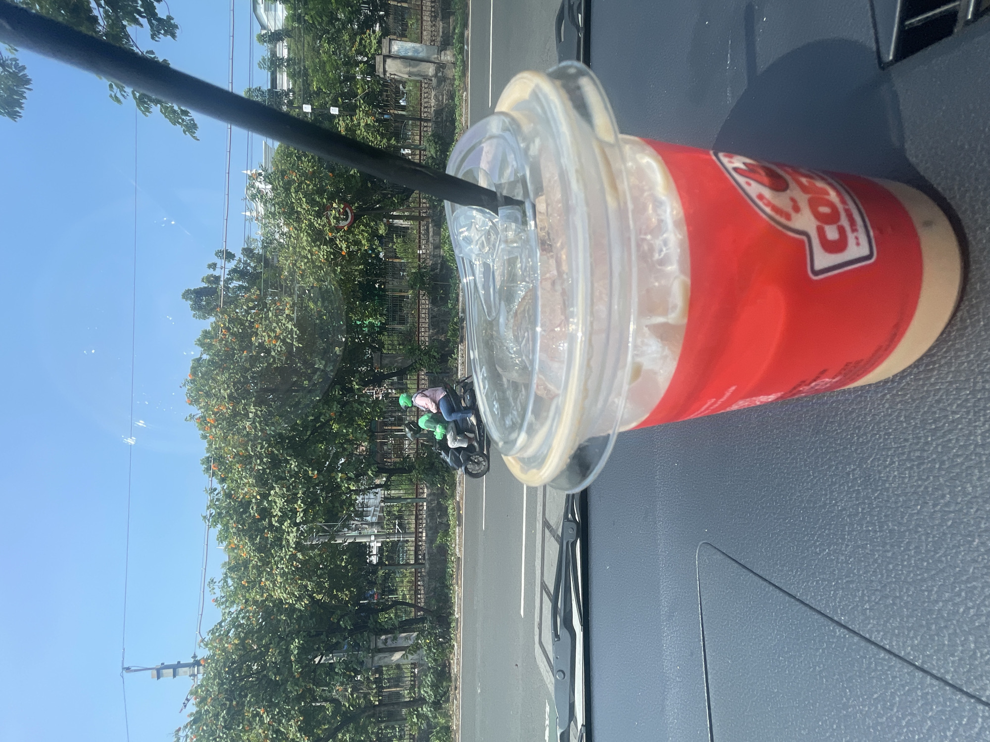 A close-up, eye-level shot focuses on a red coffee cup with a clear lid and black straw in the foreground. The cup has a logo that appears to read "COFFEE" in white lettering with a red flame graphic. Beyond the cup, a street scene unfolds. A motorcycle carrying two individuals wearing helmets, one green and one light blue, travels on the right side of the road. The road is paved and marked with white lines. Lush green trees with some orange-colored foliage line the street, and a black metal fence runs behind them. In the background, several white buildings are visible. The sky is a clear, bright blue. The image is taken from inside a vehicle, with the dashboard visible in the bottom foreground. The scene is consistent with a street in South Jakarta, Indonesia, likely during daytime due to the clear blue sky and bright lighting.